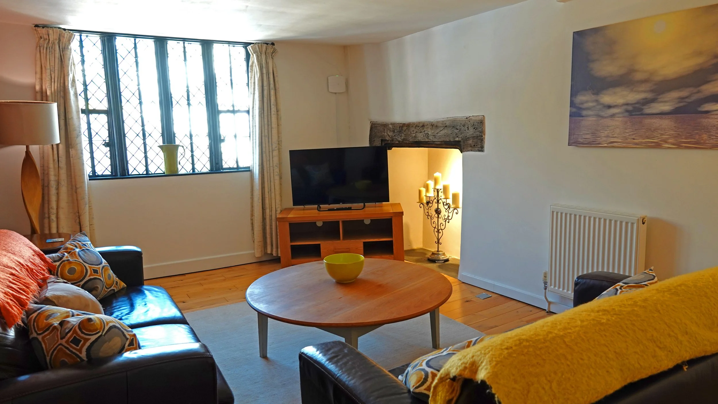 Living room with two leather sofas, a wooden coffee table with a green bowl, a wooden TV stand with a flat screen TV, a large window with curtains, a decorative candelabrum with yellow candles, a painting of a seascape, and a fireplace with a rustic wooden mantel.