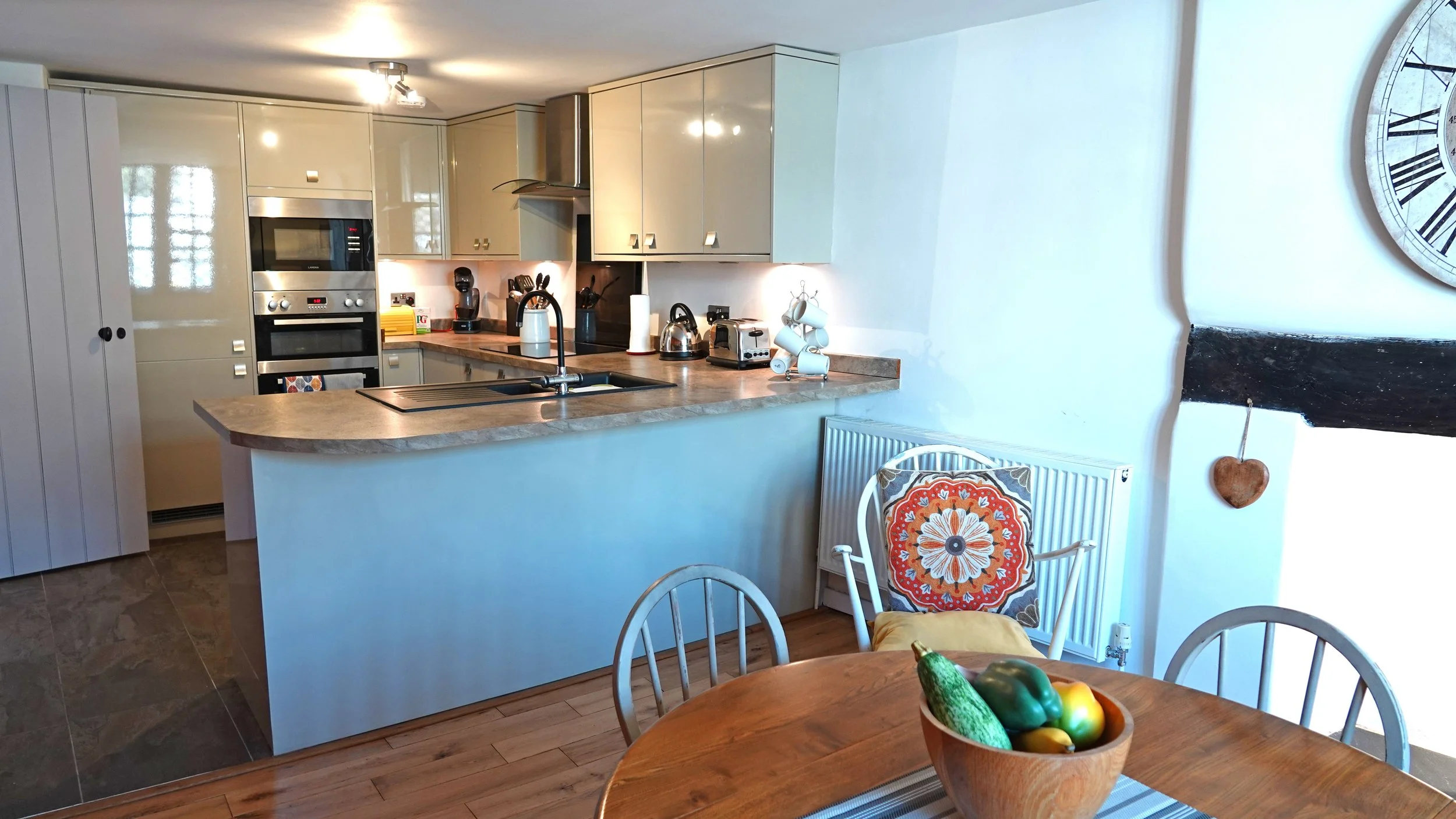 A kitchen with beige cabinets, a wooden countertop, and various appliances including a microwave, oven, coffee maker, toaster, and kettle. In front, a round wooden dining table has a bowl of vegetables, and nearby a chair has a decorative cushion. To the right, a radiator with a colorful cushion, a wall clock, and a small hanging heart decoration are visible.