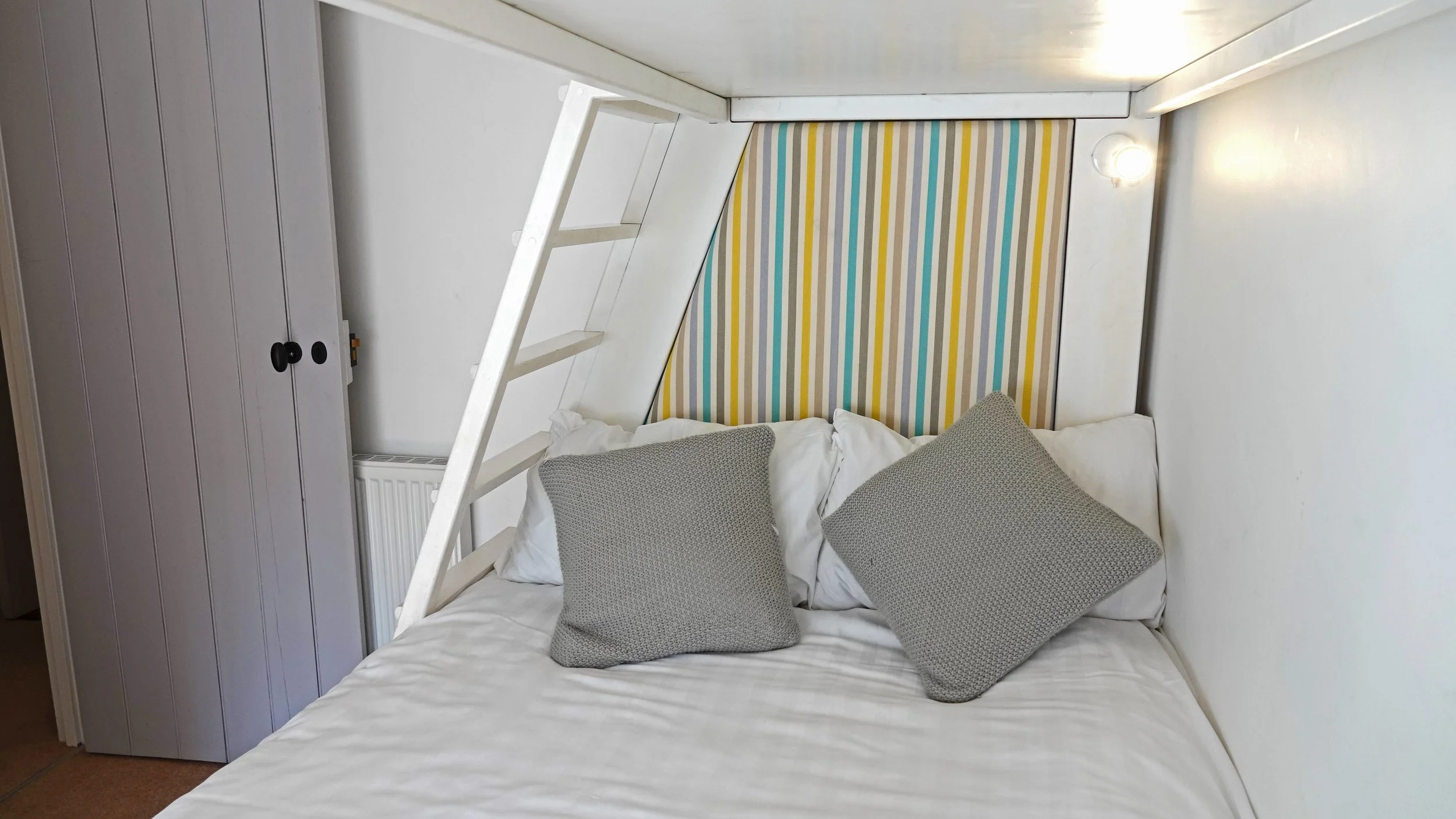 A cozy white bed with two gray pillows, a striped colorful headboard, a white ladder, a white wardrobe, and a fan, inside a small room with white-walled ceiling.