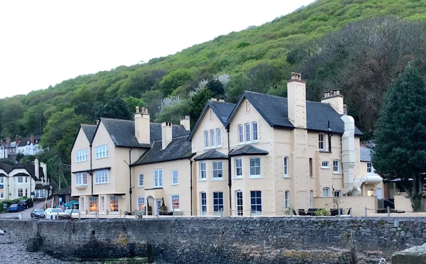 A large cream-colored mansion with multiple chimneys and black roof, situated along a stone waterfront, with lush green hillside in the background.