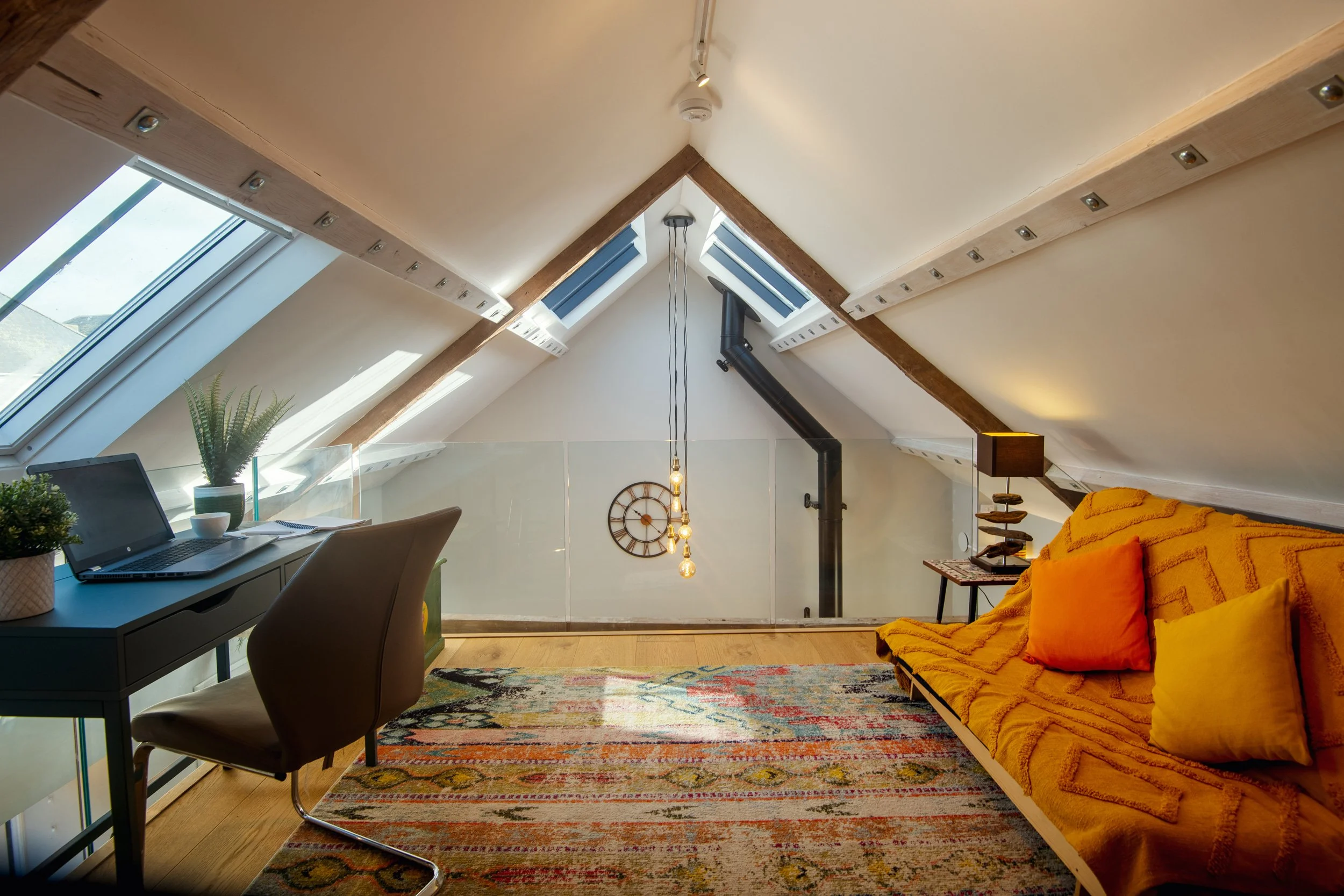 Attic room with sloped ceiling, skylight windows, a desk with a laptop and plants, a colorful rug, a yellow sofa with orange and yellow pillows, a small side table with a lamp, and a wall clock.