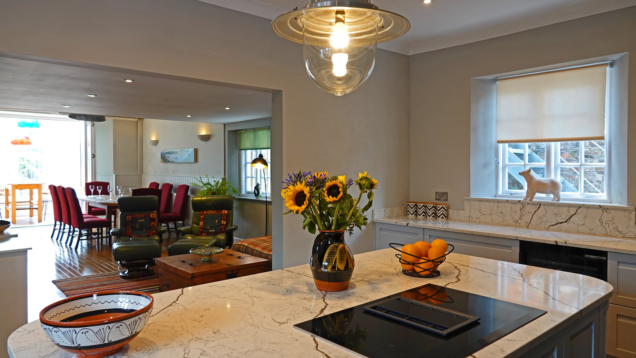 Modern kitchen with marble countertop, vase of sunflowers, bowl of oranges, window with animal figurine, and kitchen appliances, opening to a dining area with chairs and a living space.