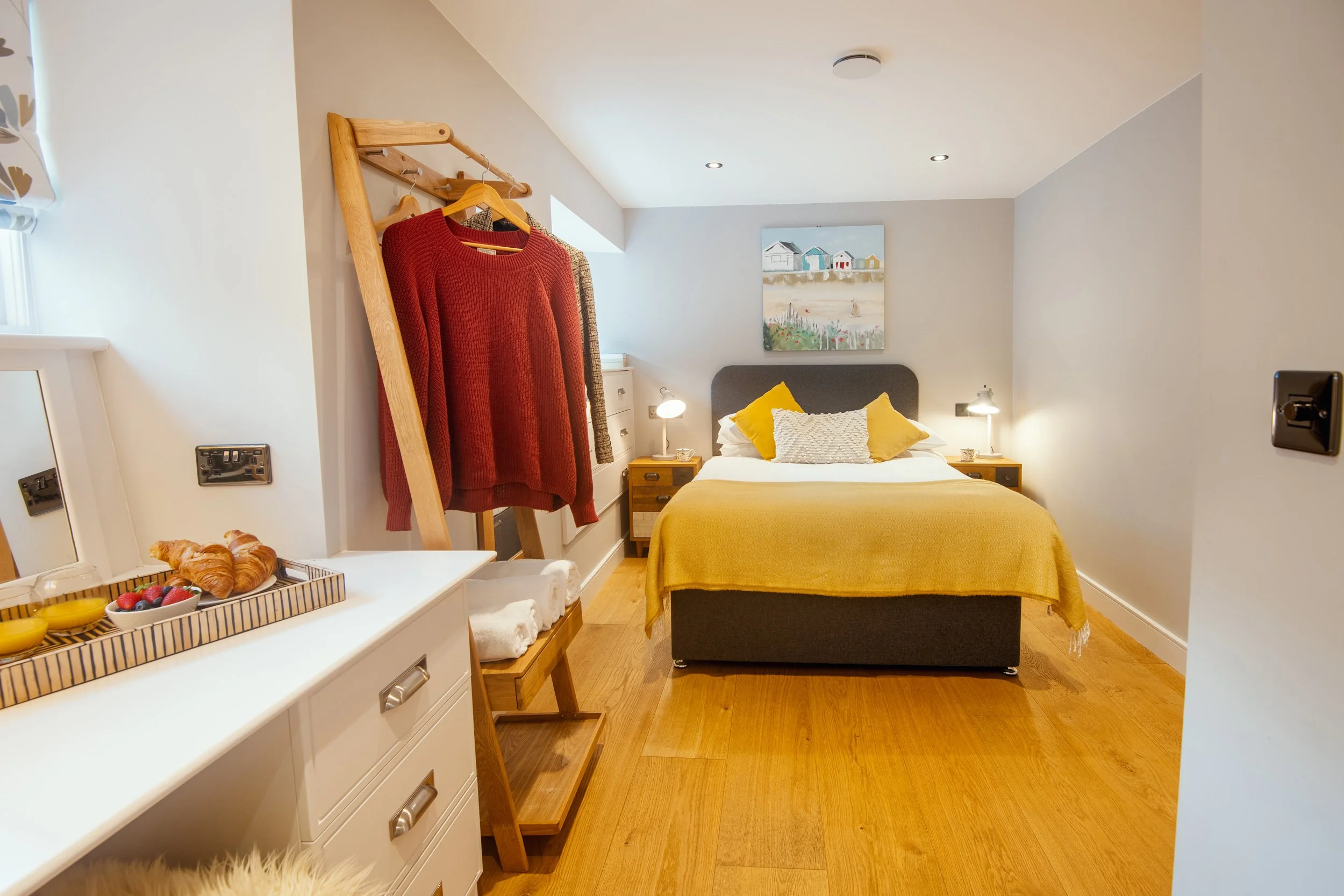 A cozy bedroom with a bed featuring a gray headboard, white bedding, and yellow pillows. Wooden nightstands with lamps are on each side of the bed, and a painting of beach huts hangs above. There is a wooden clothes rack with sweaters, a white dresser with a tray of breakfast items, and hardwood floors.