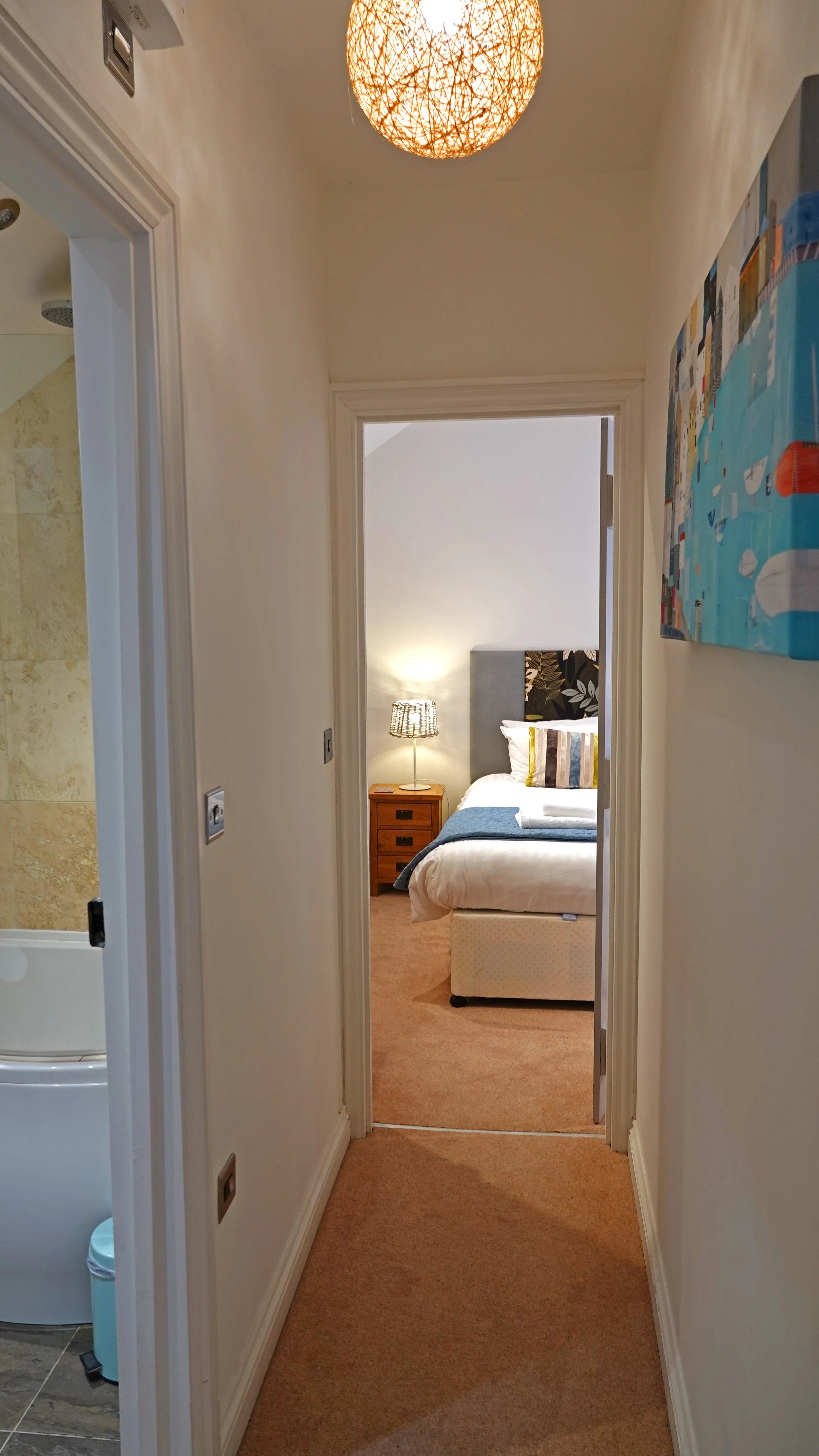 A hotel hallway leading to a bedroom with a bed, nightstand, and lamp, with a partial view of a bathroom on the left.