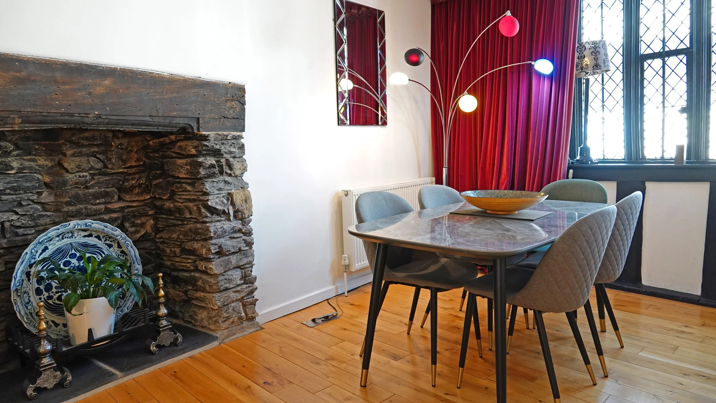 Living room with a stone fireplace on the left, a mid-century modern dining table with six chairs, a decorative bowl on the table, a multi-colored arc lamp, red curtains, and a large window with black trim and diamond-patterned grilles.