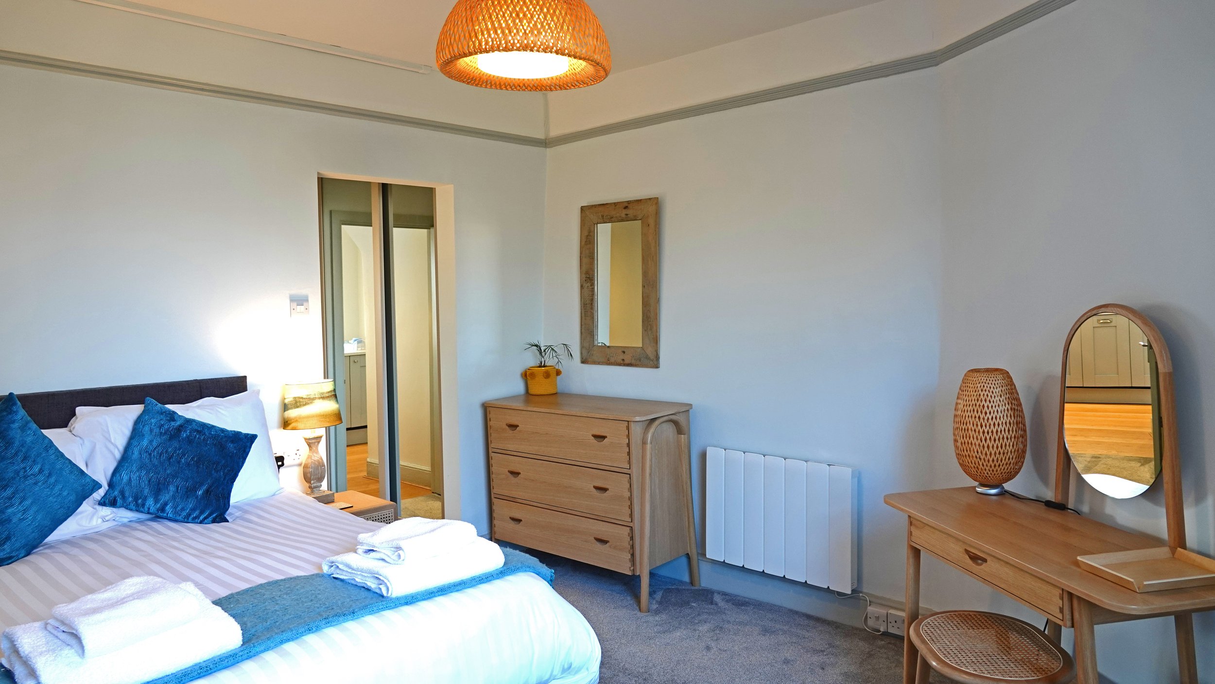 A cozy bedroom with a bed, blue pillows, white linens, a side table with a lamp, a wooden dresser with a plant, a mirror, and a wooden vanity with a lamp, in a well-lit room.