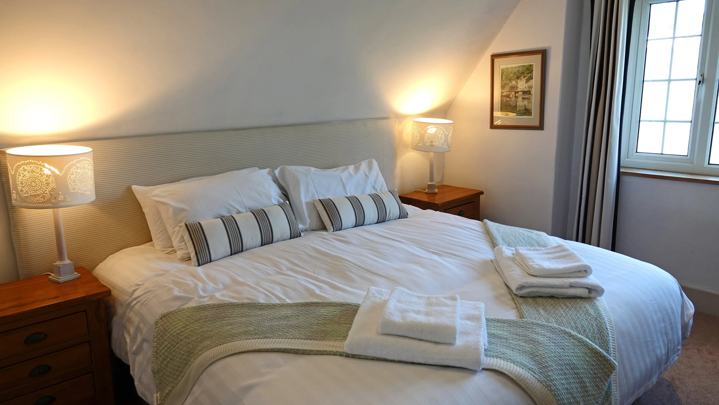 A neatly made bed with white linens, striped pillows, and folded towels on top, flanked by nightstands with table lamps, in a cozy bedroom with a window with curtains and a framed painting on the wall.