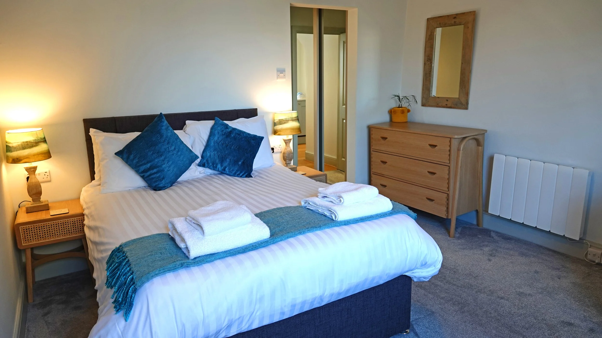 A neatly made bed with white linens and blue pillows in a cozy bedroom. There are two sets of folded white towels on the bed, with a teal throw blanket. The room features wooden furniture, including two bedside tables with lamps, a dresser with a plant on top, a mirror on the wall, and a radiator near the floor.