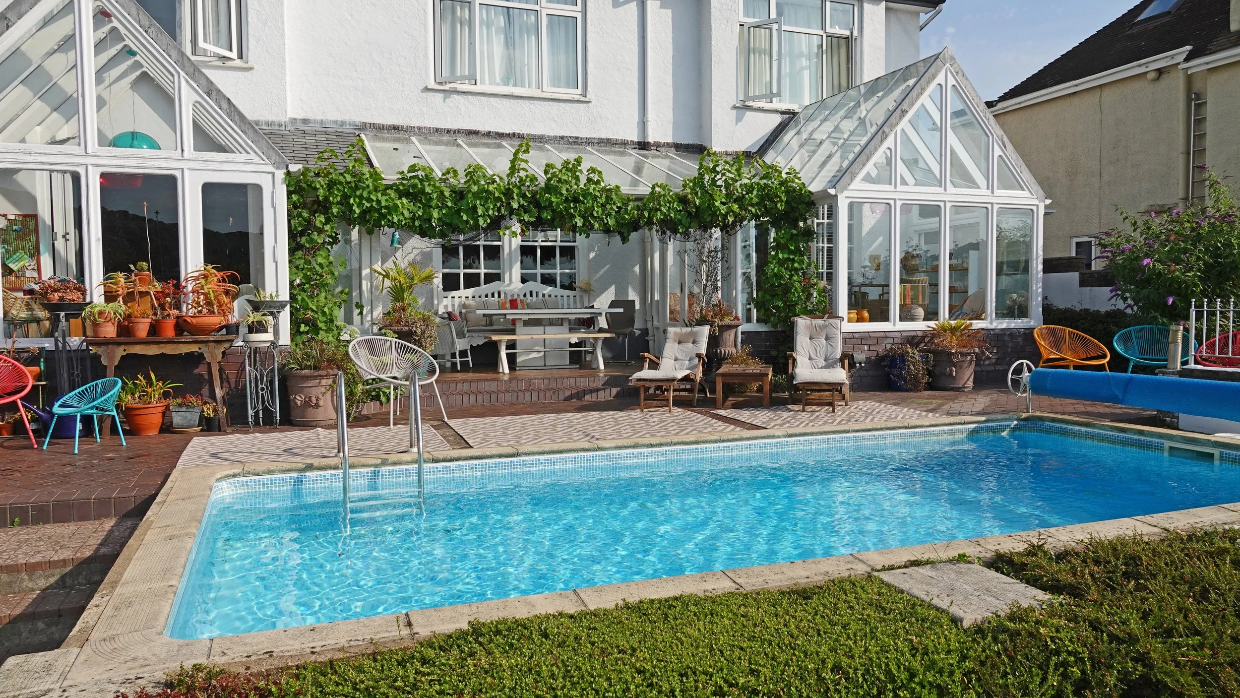 A backyard with a swimming pool in front of a white house with a glass greenhouse structure. There are various chairs and tables, plants, and a trellis with green vines.