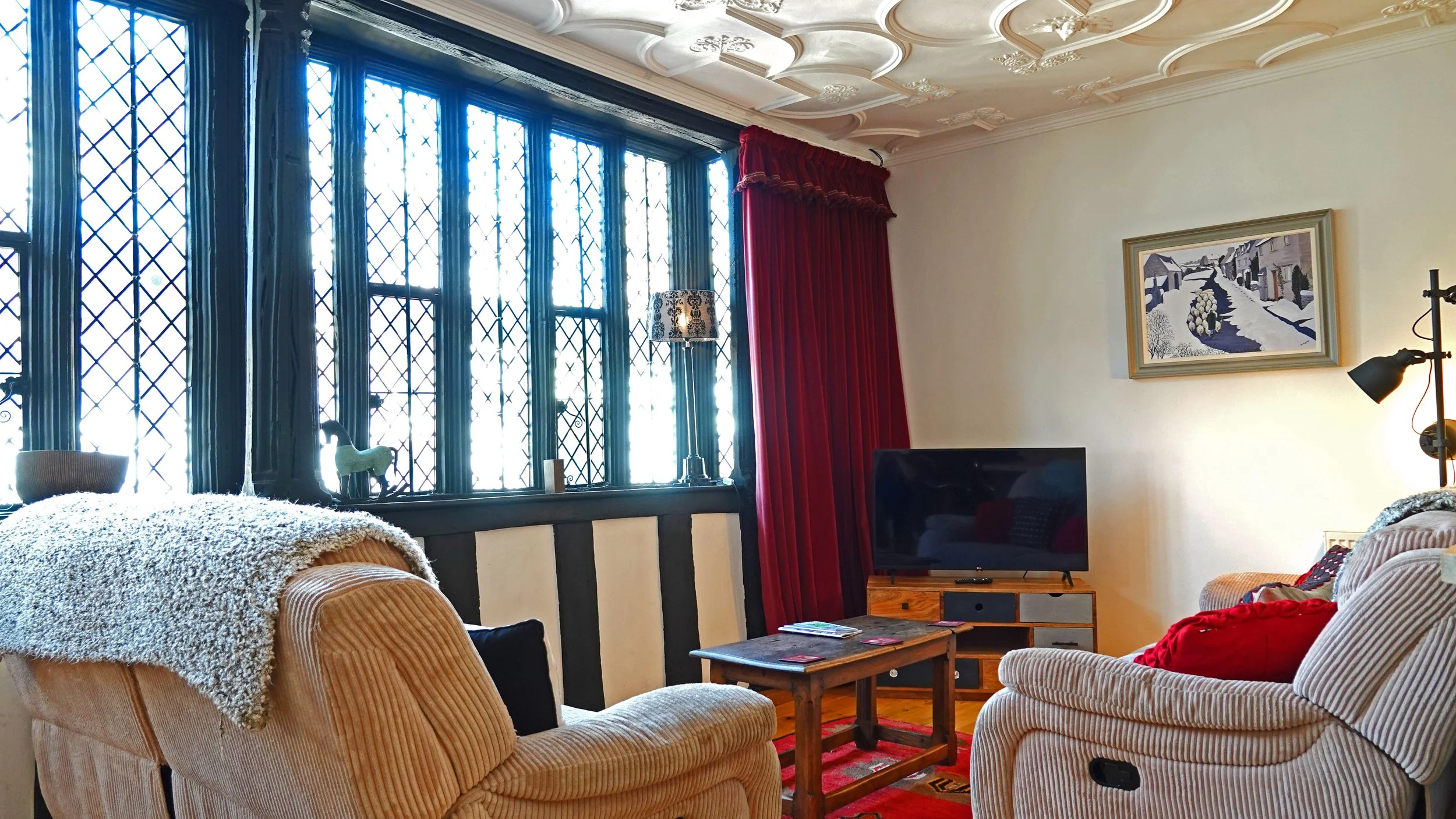 Living room with patterned ceiling, large leaded glass window with black trim, red velvet curtain, beige striped armchair, orange striped armchair with red pillow, flat-screen TV on wooden stand, wooden coffee table with magazines, wall art depicting snowy cottages, standing lamp, and decorative items on window sill.