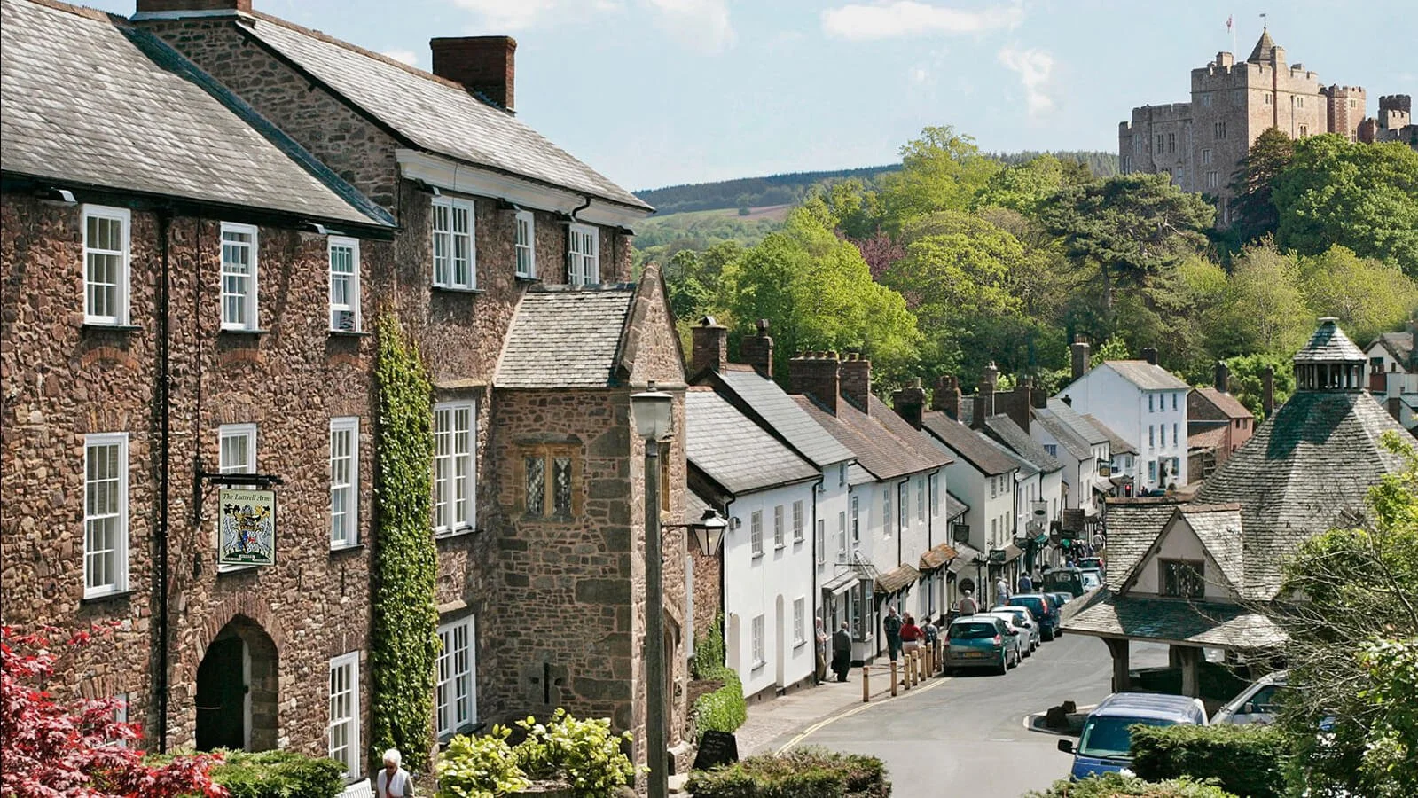 Hilly street in a rural village with stone and white houses, cars parked along the road, people walking, and greenery including trees and bushes, with a historic castle in the background.