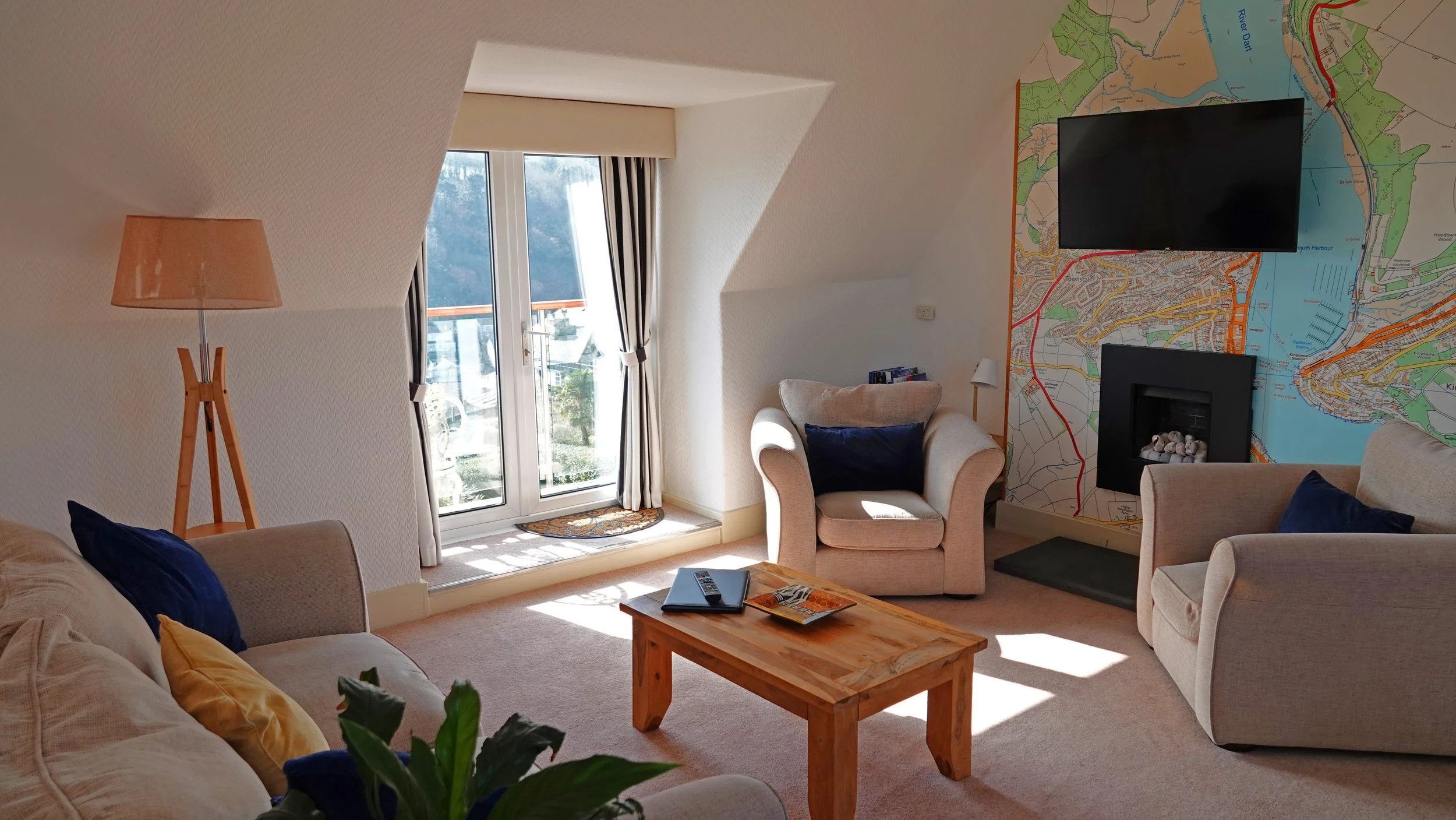 Living room with beige couches, a glass door leading outside, a wooden coffee table with magazines, a wall decorated with a colorful map, a wall-mounted TV, a fireplace, a beige armchair, a floor lamp, and velvet pillows in navy blue and mustard yellow.
