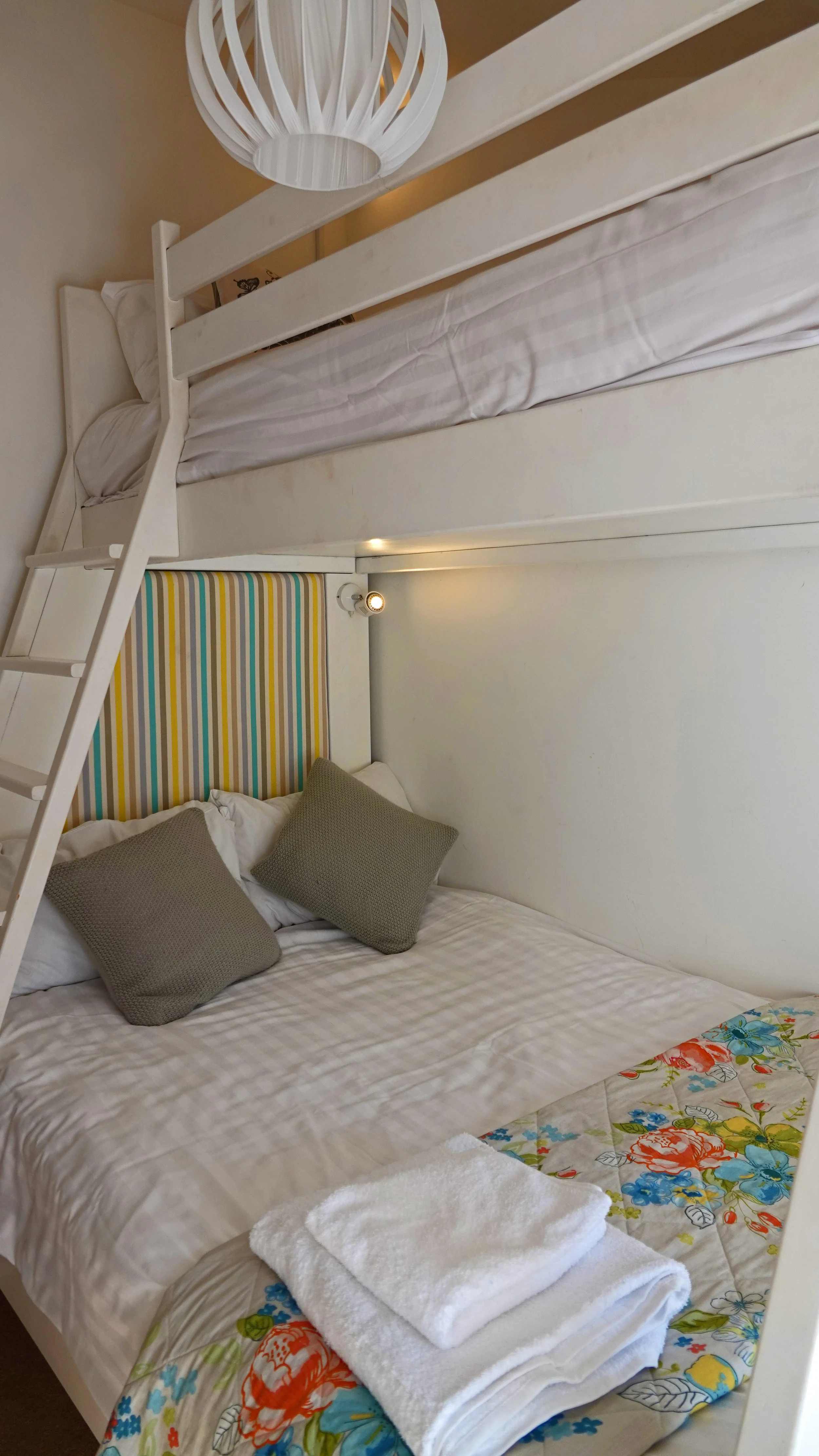 A bedroom with a bunk bed, the top bunk has white bedding and the lower bunk has a floral bedspread with two gray pillows and folded white towels.