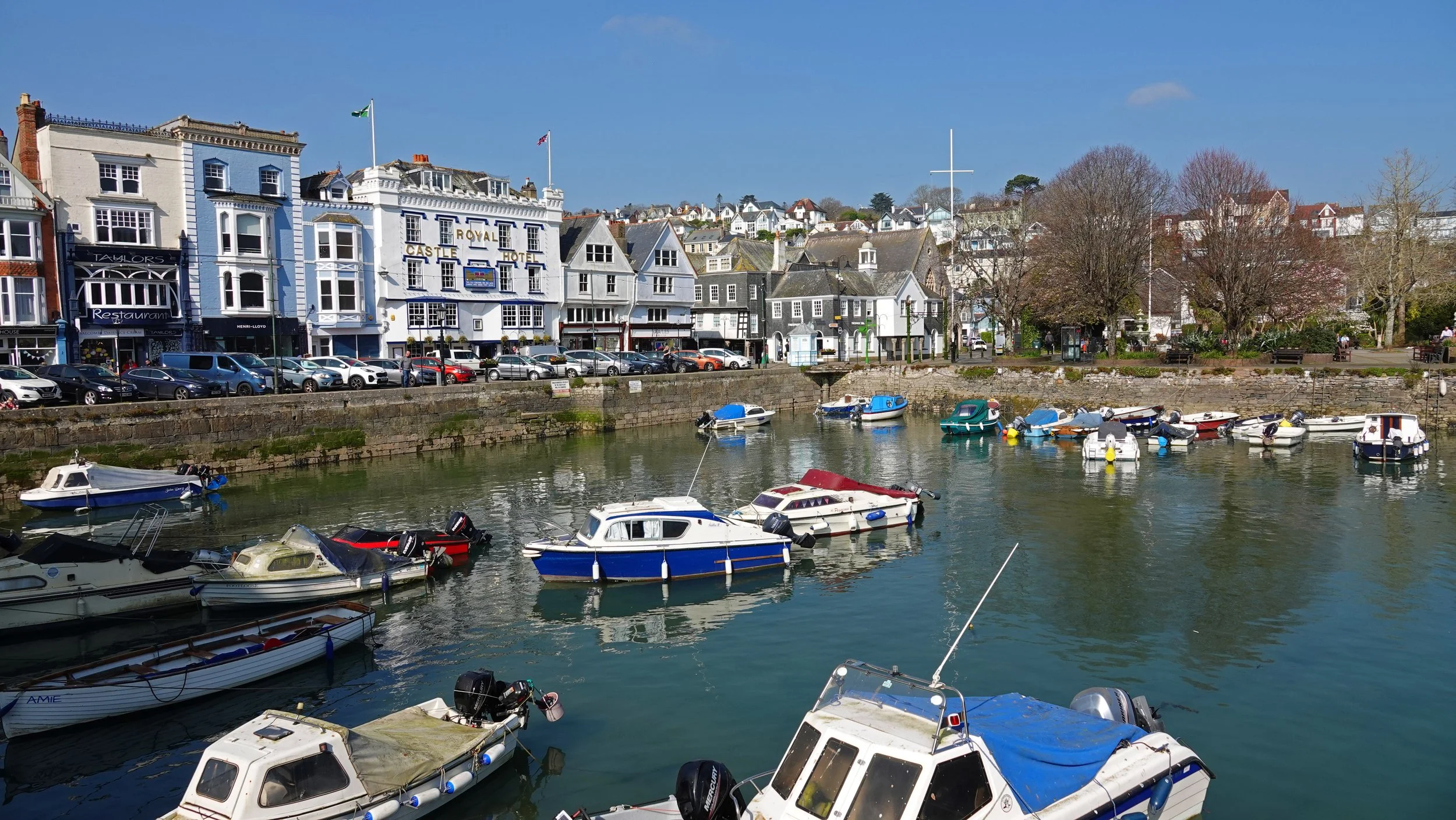 Colorful boats docked in a harbor with buildings and trees along the waterfront under a clear blue sky.