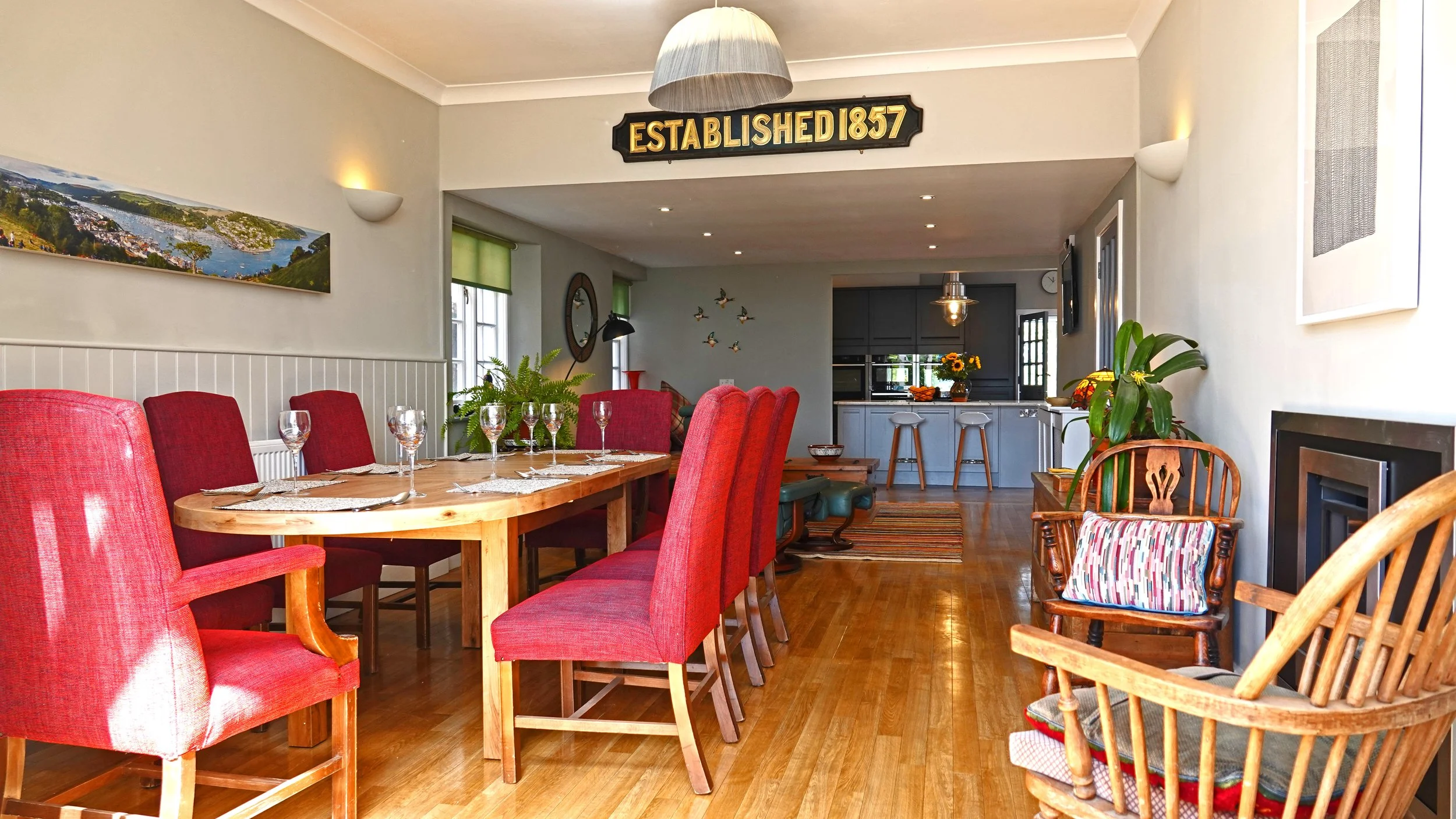 A dining area with a large wooden table surrounded by red upholstered chairs. The room has hardwood floors, a painting on the wall, and a sign that reads 'ESTABLISHED 1857' hanging above the doorway leading to a kitchen with dark cabinets and a breakfast bar with three barstools.
