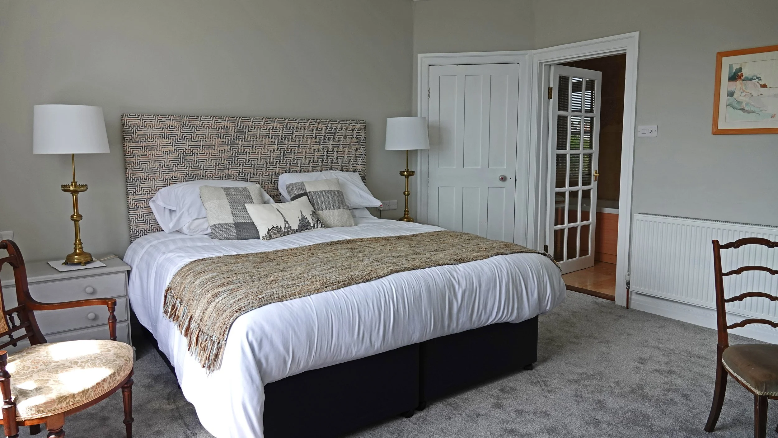 A neatly made bed with a patterned headboard, decorative pillows, and a throw blanket, flanked by two brass lamps on white nightstands, in a bedroom with gray walls, carpeted floor, and framed art on the wall.