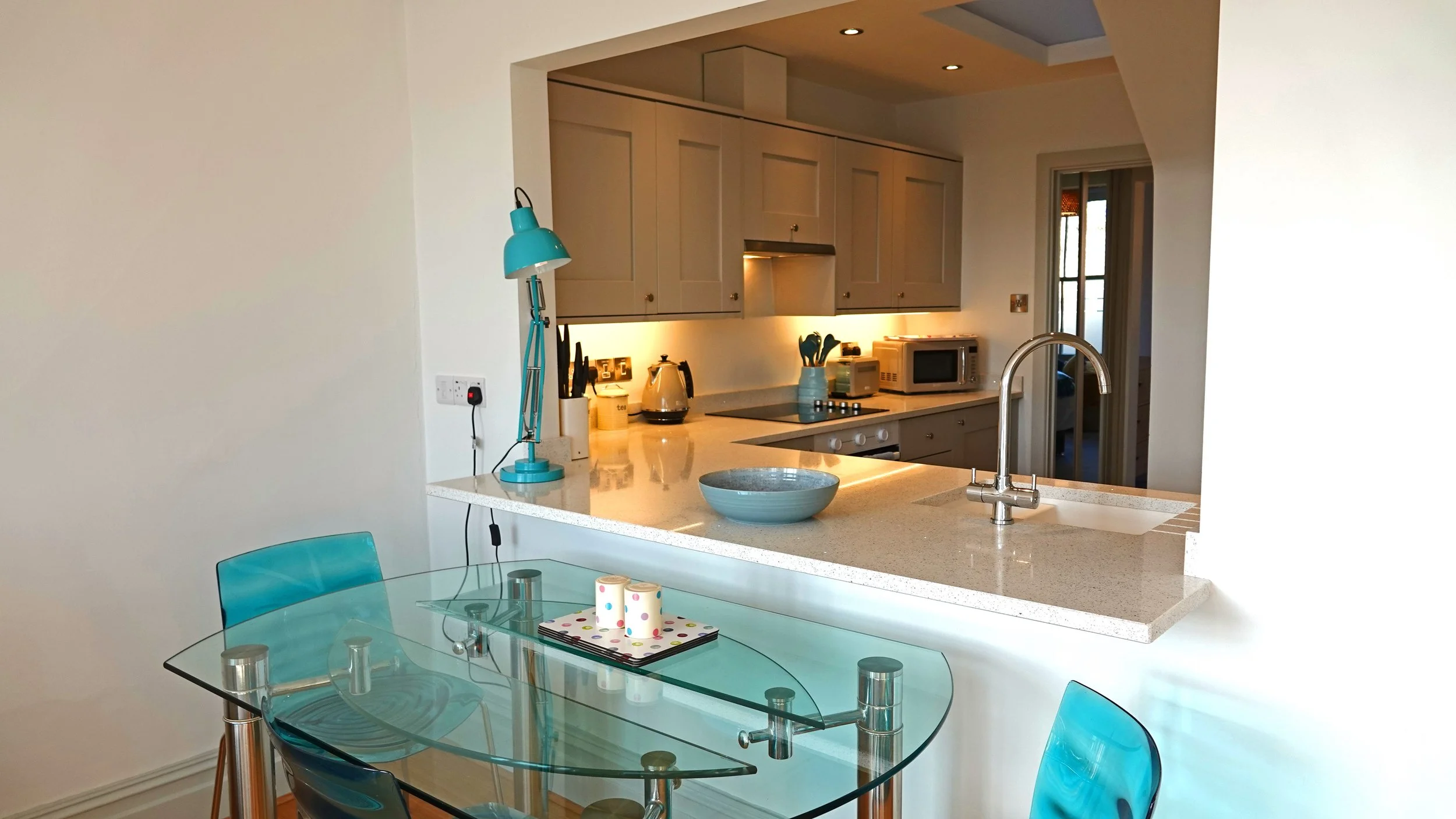 Modern kitchen with beige cabinets, microwave, kettle, and blue decor accents, visible through a breakfast bar with a bowl on the counter and a glass dining table with blue chairs in the foreground.