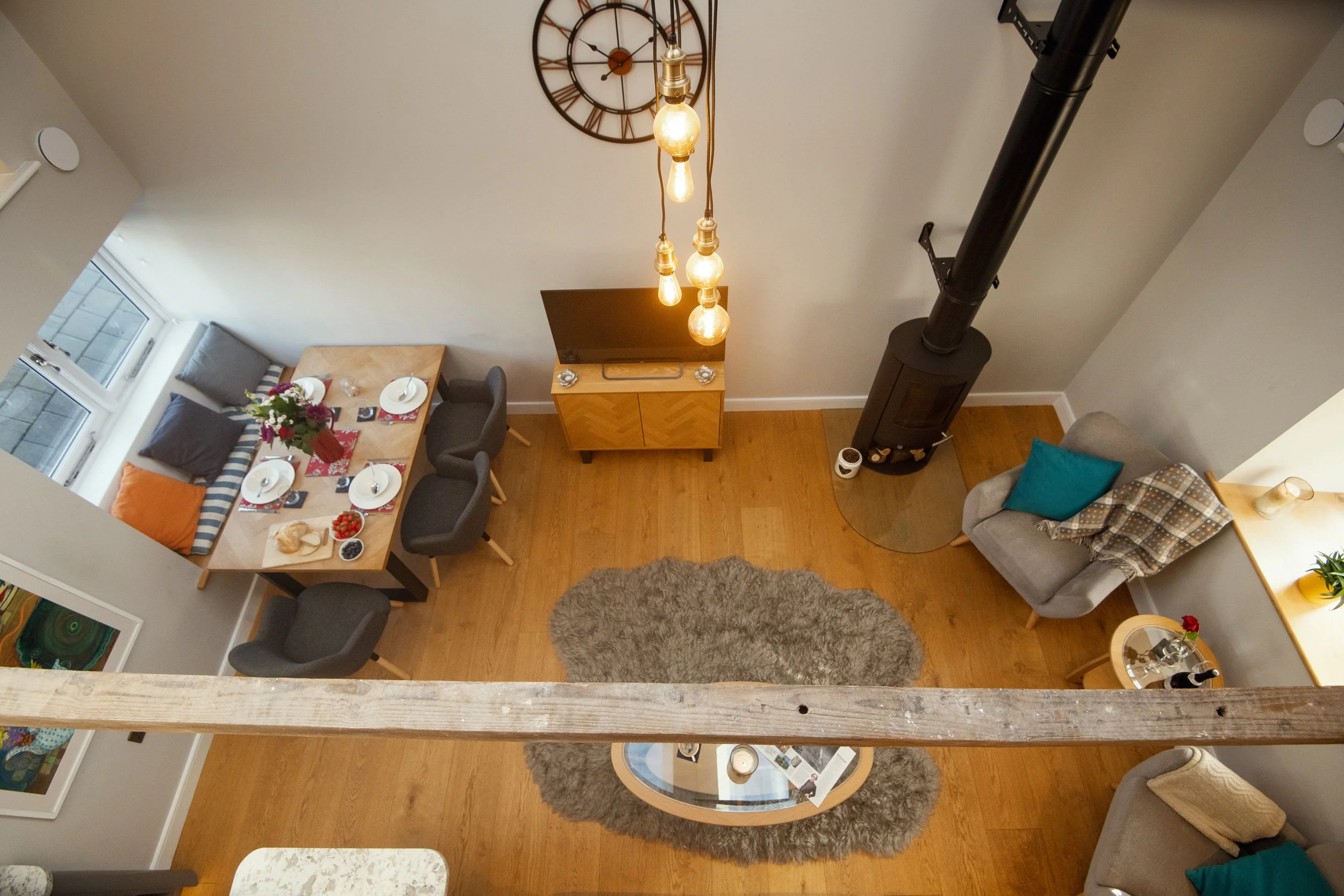 Overhead view of a cozy living and dining area with a wooden dining table set for four, a plush gray area rug, a round glass coffee table, a wood stove, a TV on a wooden stand, and seating including a gray armchair with teal pillow and a beige armchair with a blue pillow, sunlight streaming through windows, and various decor items.