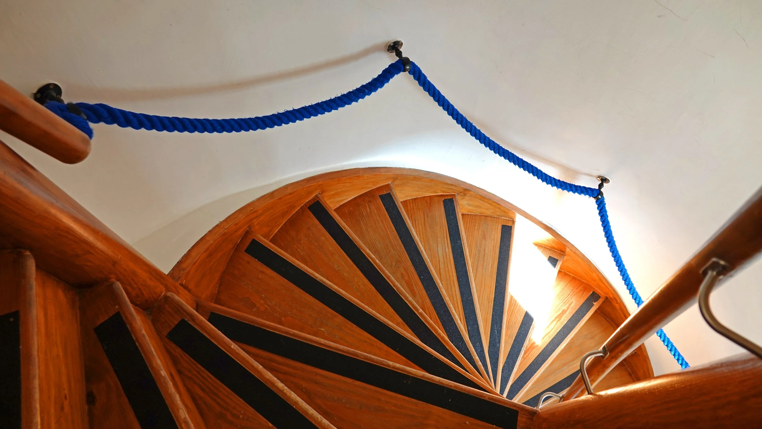 Top view of a wooden spiral staircase with black anti-slip strips on each step and a blue braided safety rope along the railing.