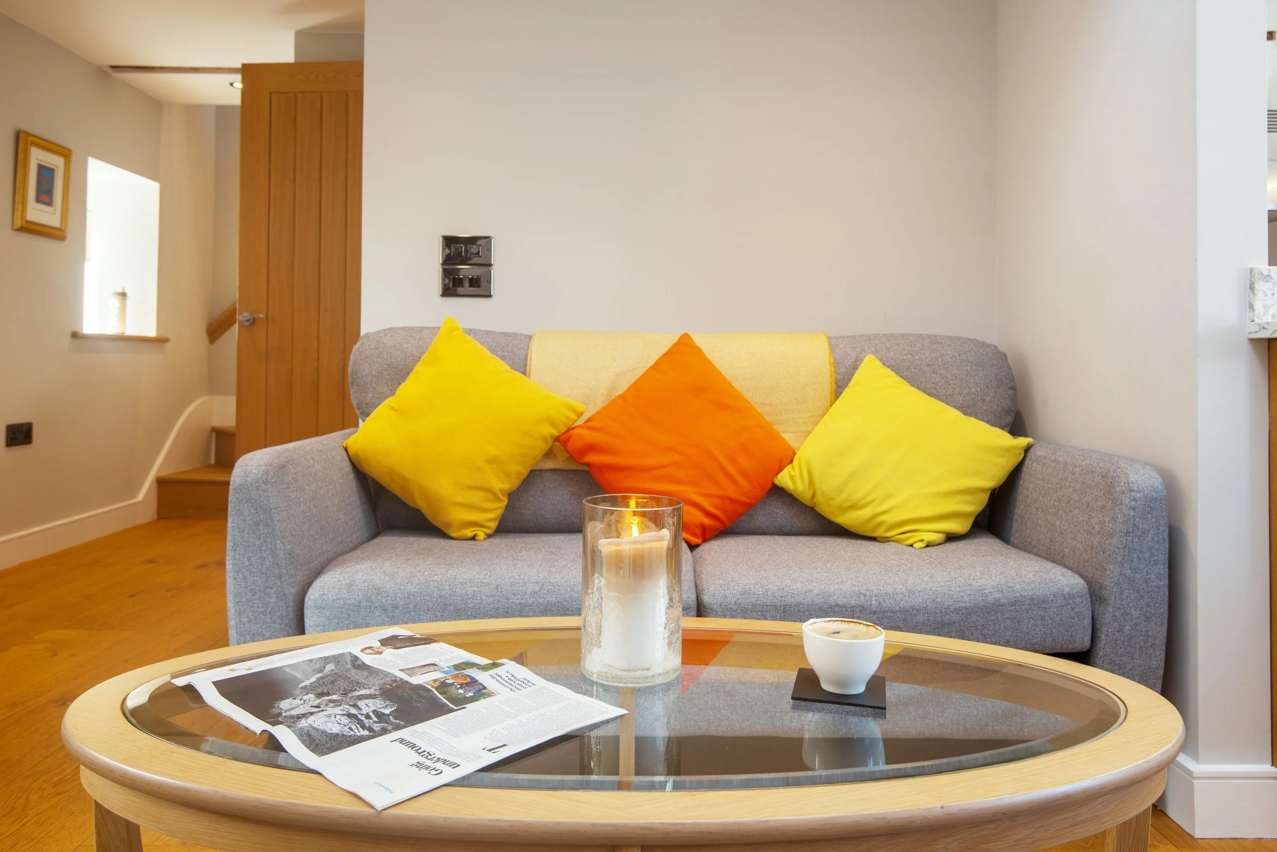 Living room with gray couch, yellow and orange pillows, a glass-top coffee table with a candle, a newspaper, and a cup of coffee.