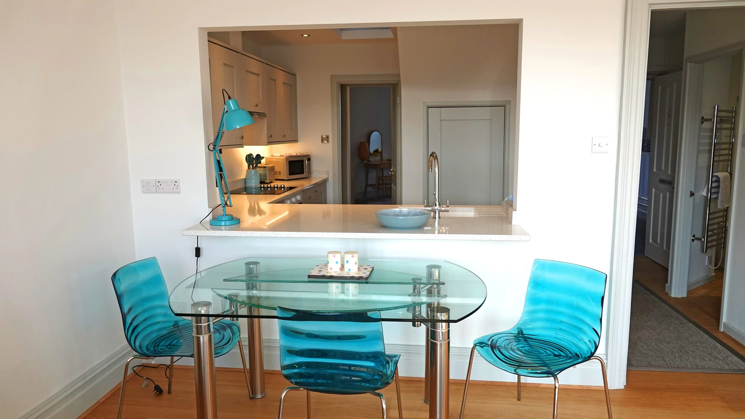 A modern kitchen and dining area with a glass dining table and four blue translucent chairs. The kitchen has beige cabinets, a microwave, and a teal desk lamp on the counter. There are candles on the table, and the kitchen has an open view to a hallway and living space.