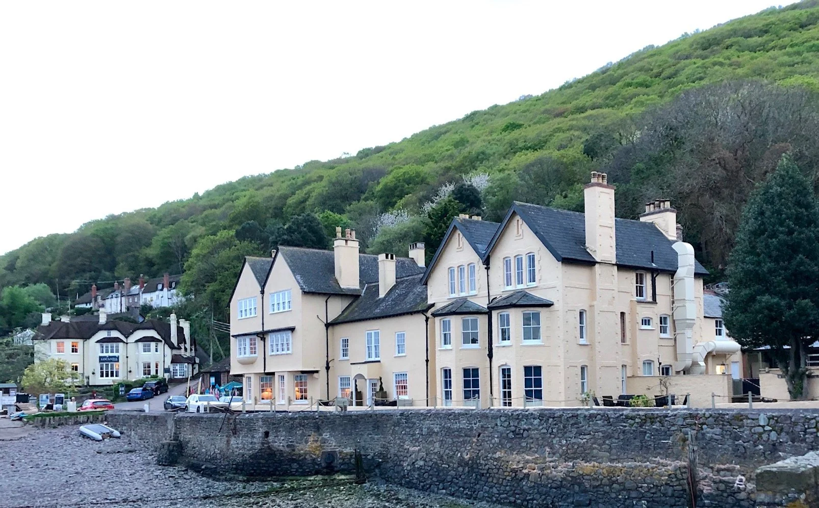 A row of cream-colored buildings with dark roofs and chimneys, situated along a rocky waterfront, with green forested hills in the background.