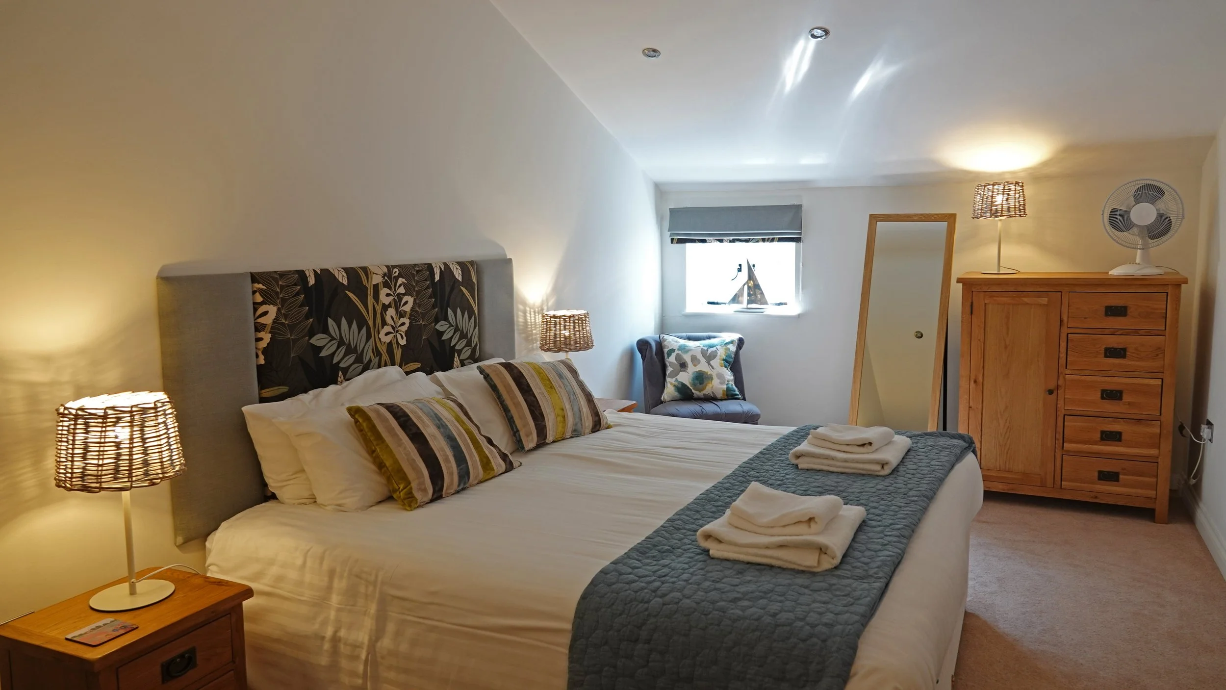 Cozy bedroom with a bed featuring a patterned headboard, striped pillows, and towels on top. The room has a wooden dresser with a fan and lamp, a chair with a cushion, small window, mirror, and bedside lamp.