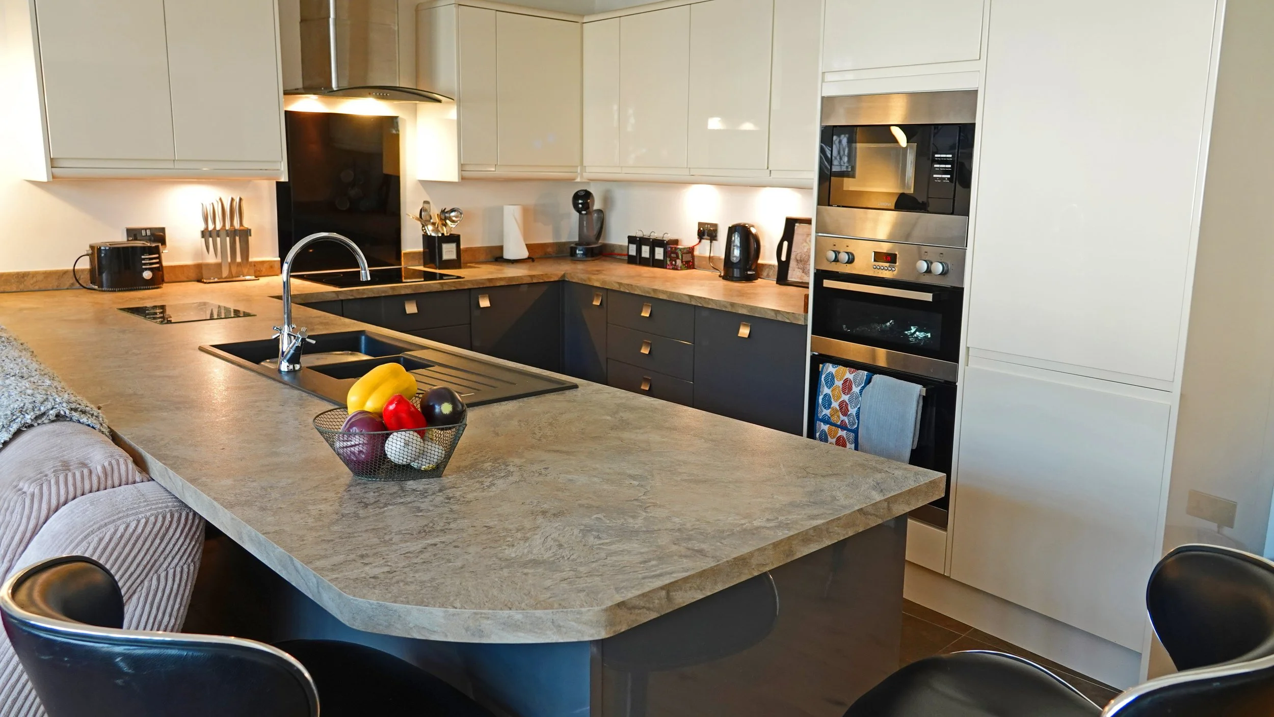 Modern kitchen with beige cabinets, black lower cabinets, and a beige marble countertop. Contains a sink, fruit basket, black stove, microwave, oven, toaster, and various small appliances.