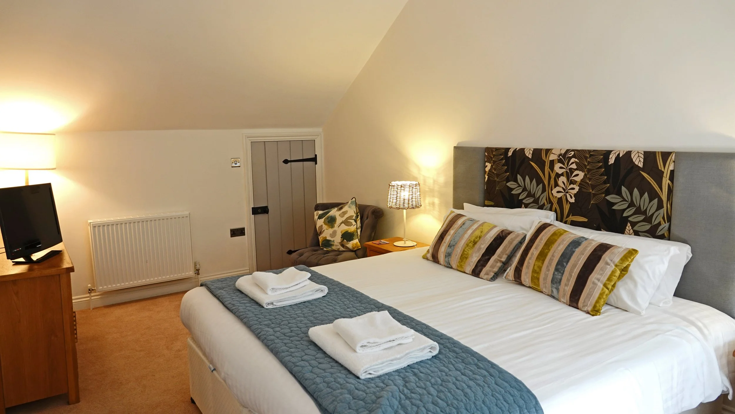 A hotel room with a large bed, striped pillows, a headboard with a floral pattern, a bedside lamp, a TV on a wooden stand, a small armchair with a pillow, and a door.