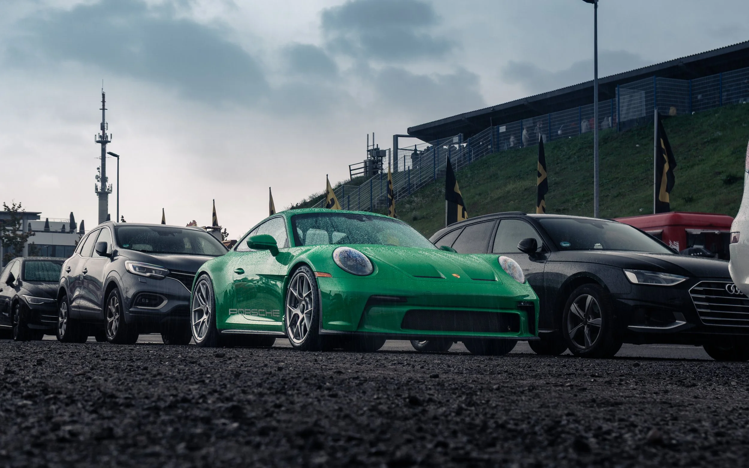Mehrere Autos, einschließlich eines grünen Porsche, sind auf einem nassen Parkplatz bei Regen, im Hintergrund Banner und ein Hügel mit Gebäuden und einem Turm.