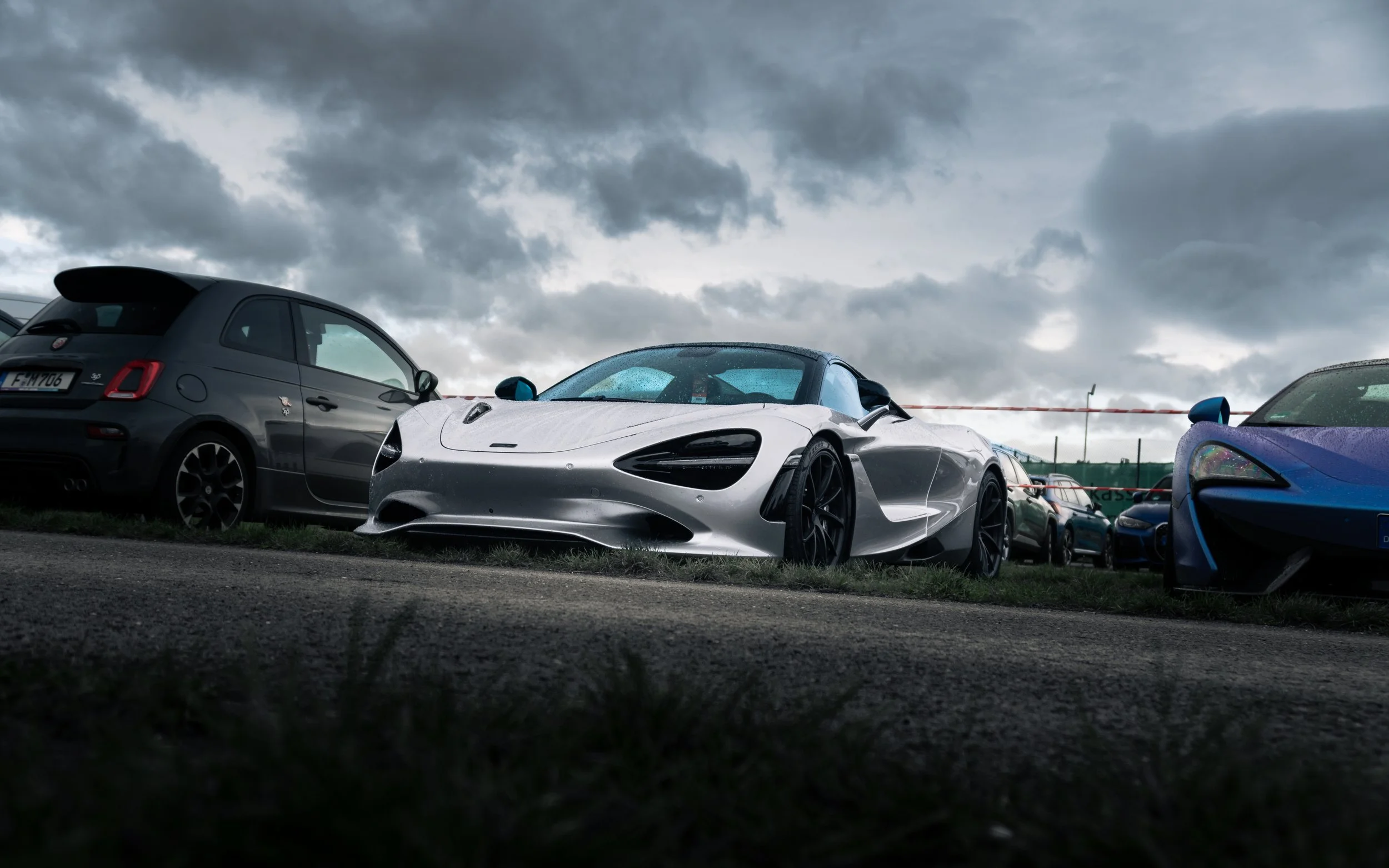 Silberner Sportwagen steht auf einem Parkplatz, umgeben von anderen Autos, bei dunklem Himmel.