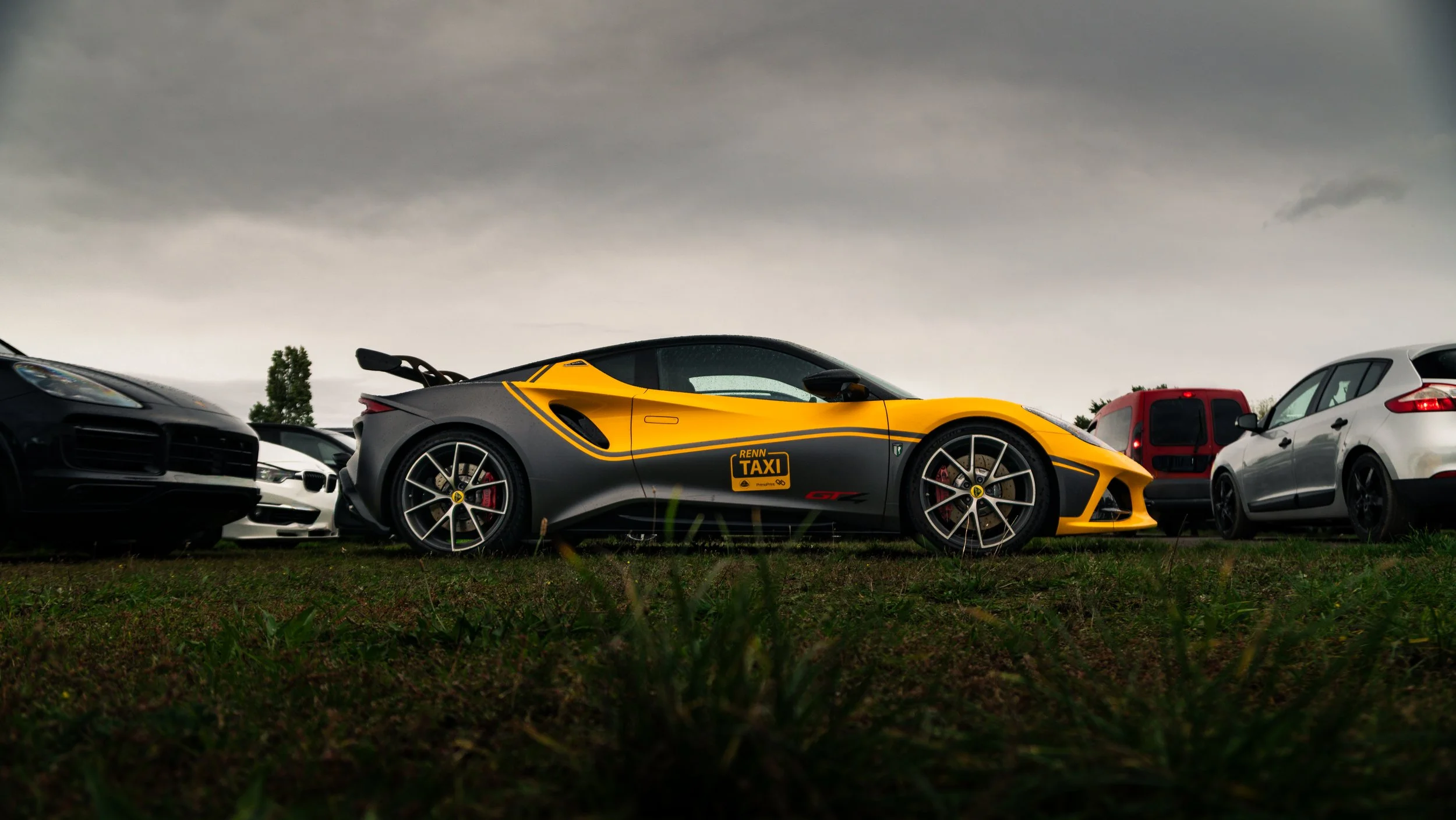Ein gelb-schwarzer Rennsportwagen, der als Taxi gekennzeichnet ist, ist in einem Parkplatz mit anderen Autos unter einem bewölkten Himmel geparkt.