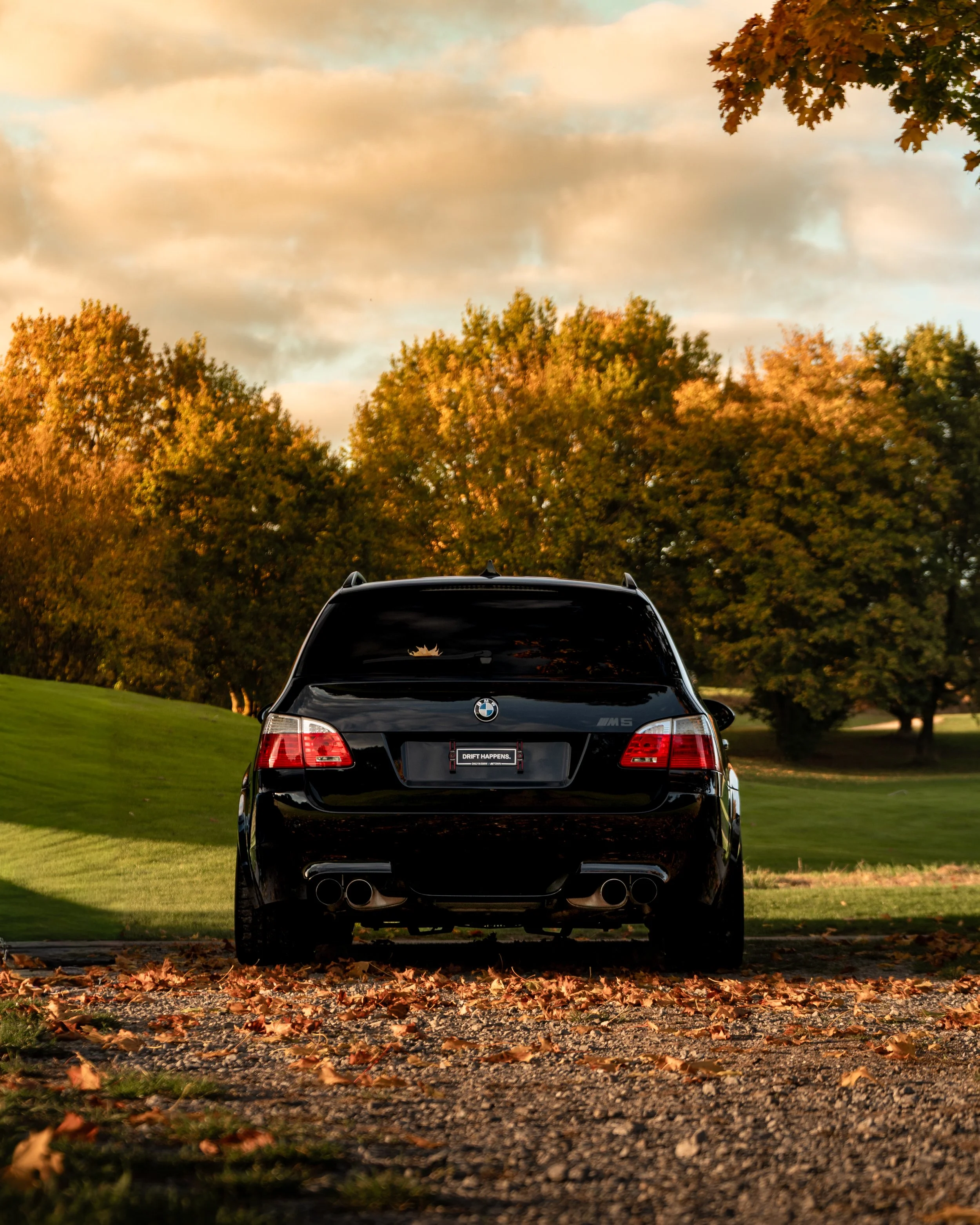 Schwarzer BMW M5 sportwagen steht auf einem Kiesweg, umgeben von Laub im Herbst, mit Bäumen und einem bewölkten Himmel im Hintergrund.