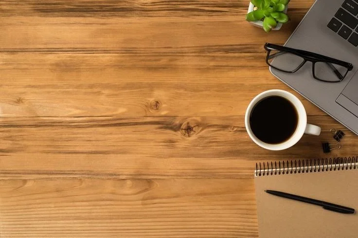 Wooden desk with a laptop, glasses, a small green plant, a cup of black coffee, a notebook, a pen, and some binder clips.