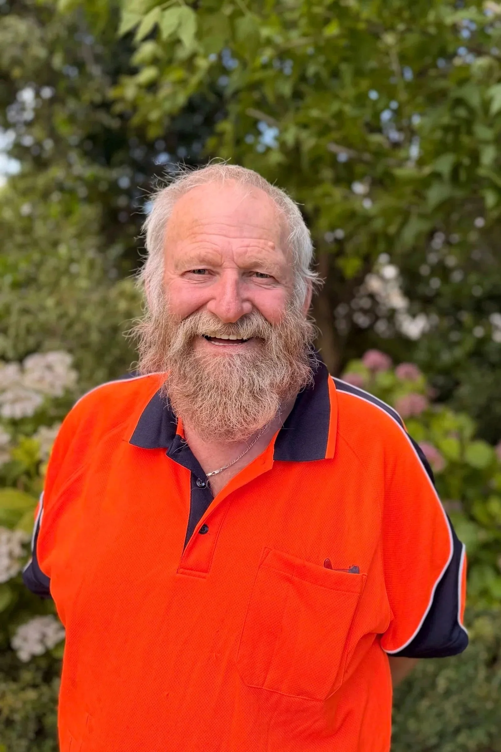 A smiling elderly man with a beard and gray hair standing outdoors, wearing an orange and black collared shirt, with a blurred green leafy background.