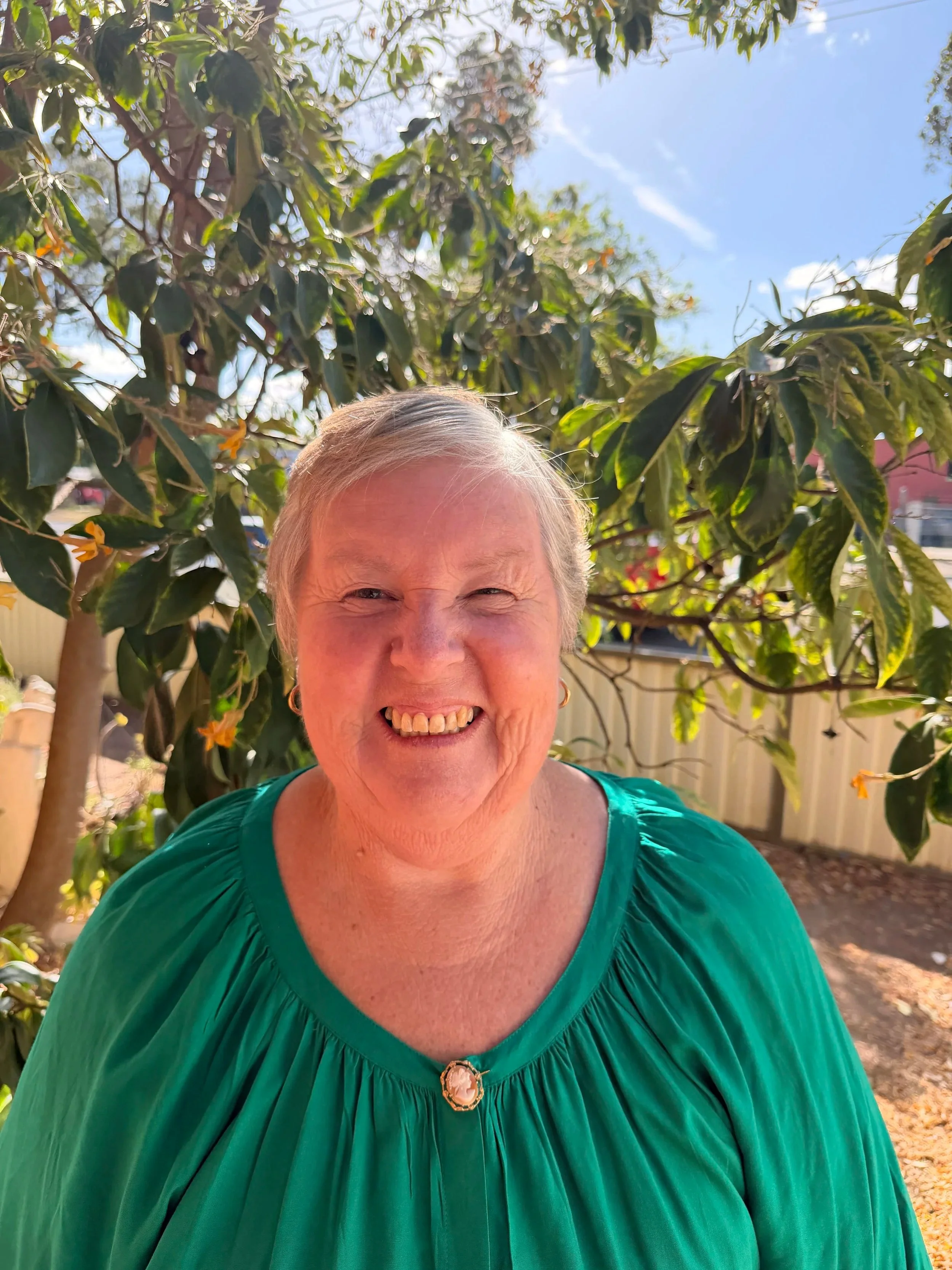 A smiling elderly woman with short gray hair, wearing a green pleated blouse with a decorative brooch at the neckline, standing outdoors in front of a tree with green leaves and orange flowers, under a blue sky.