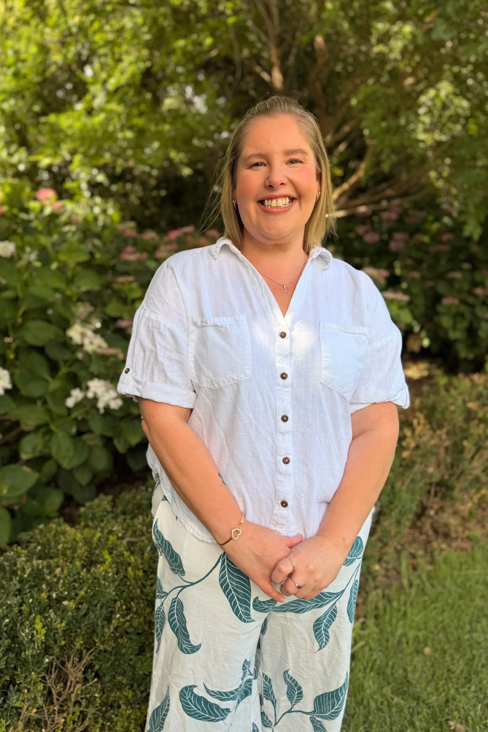 A woman with shoulder-length blonde hair, smiling and wearing a white short-sleeve button-up shirt and white pants with green leaf patterns, standing outdoors in a garden with green foliage and pink and white flowers.