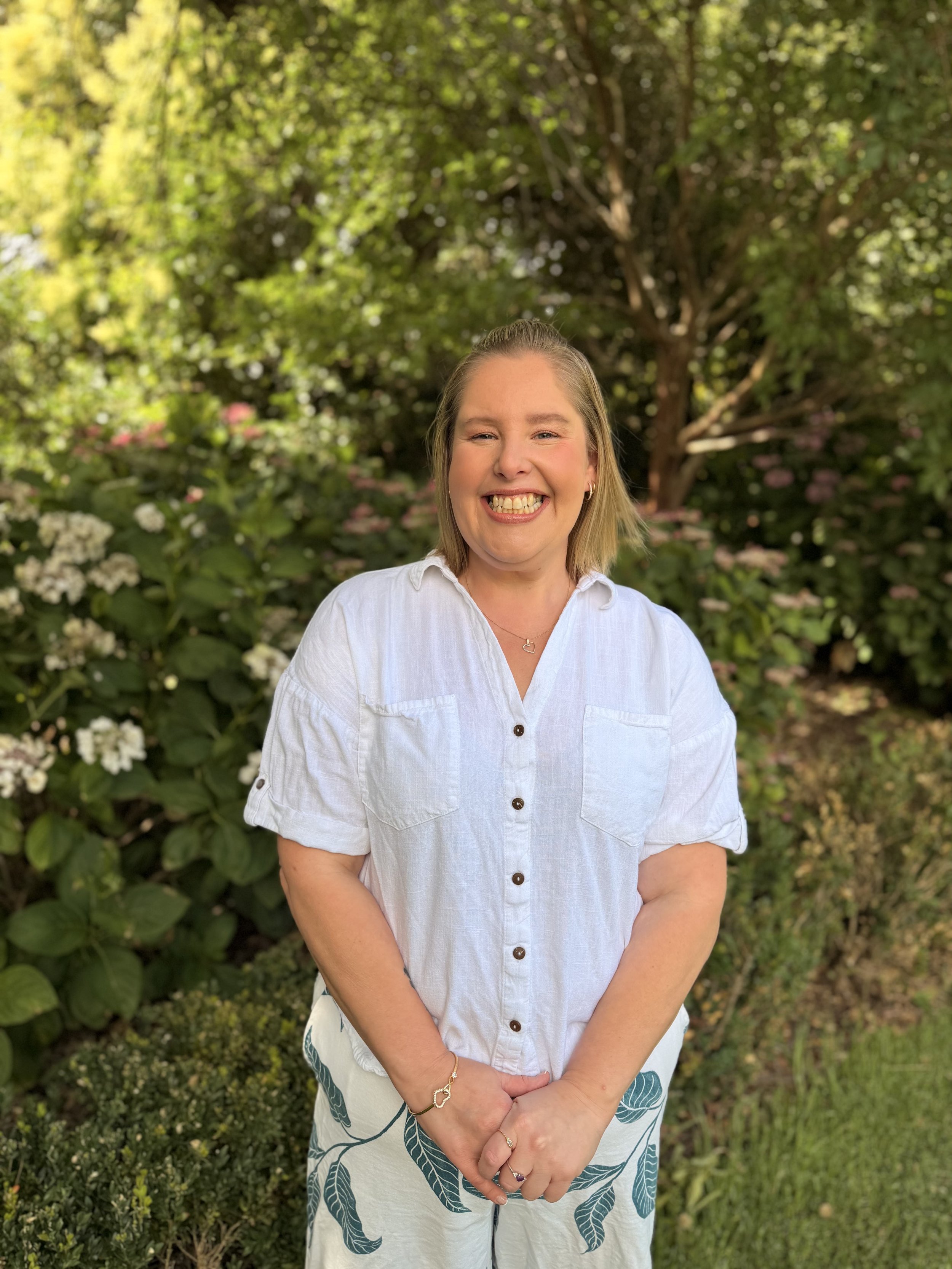 A smiling woman with shoulder-length blonde hair, wearing a white button-up shirt and patterned pants, standing outdoors in a garden with green foliage and blooming flowers.