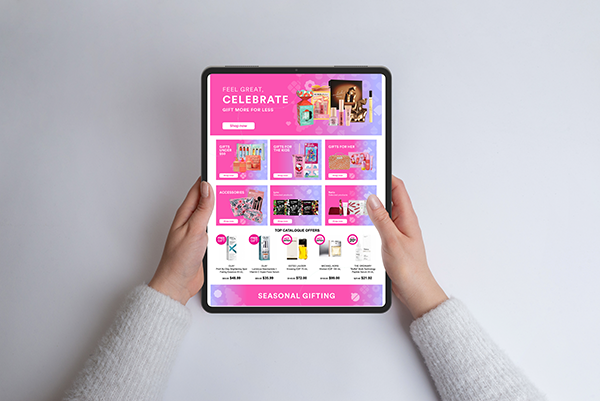 Person holding a tablet displaying a holiday gift catalog with pink and purple themes on a white wall background.