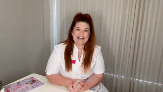 A woman with long reddish hair smiling and sitting at a table in a room with vertical blinds.