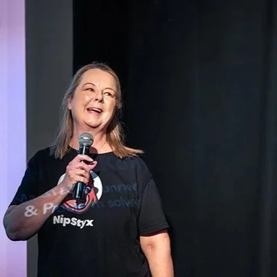 Jo Kelly smiling and holding a microphone on stage, wearing a black NipStyx T-shirt in front of a black curtain.