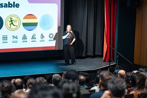 Jo Kelly at NipStyx presentation on a stage in front of a projection screen with a colorful logo and text, in front of an audience seated in a theater or auditorium.
