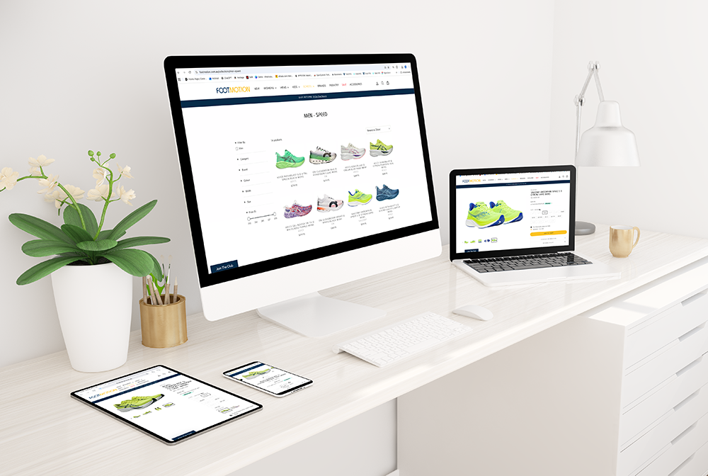 A white desk with an iMac, MacBook, iPad, and iPhone displaying an online shoe store. There is a white plant in a white pot, a small gold container with pens, a beige mug, and a white desk lamp.