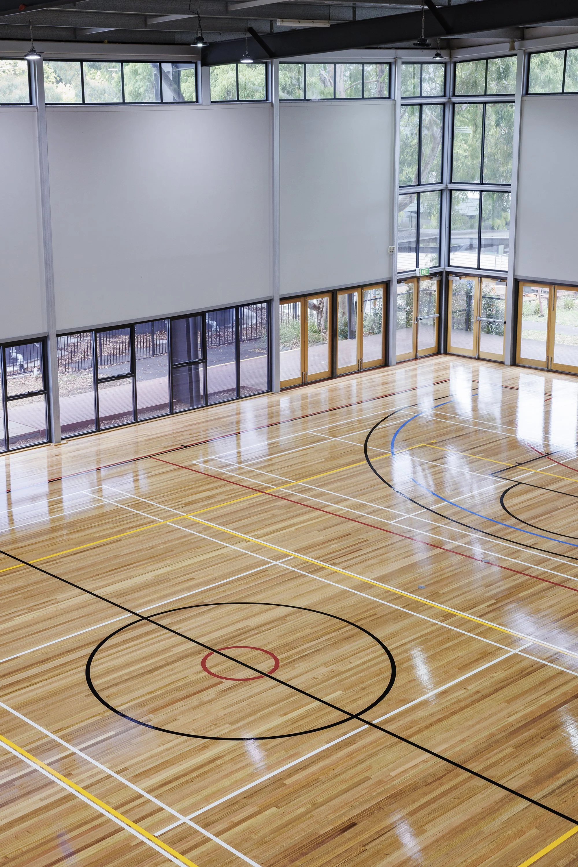 Indoor gymnasium with polished wooden floor, marked for basketball and other sports, surrounded by large windows and glass doors.
