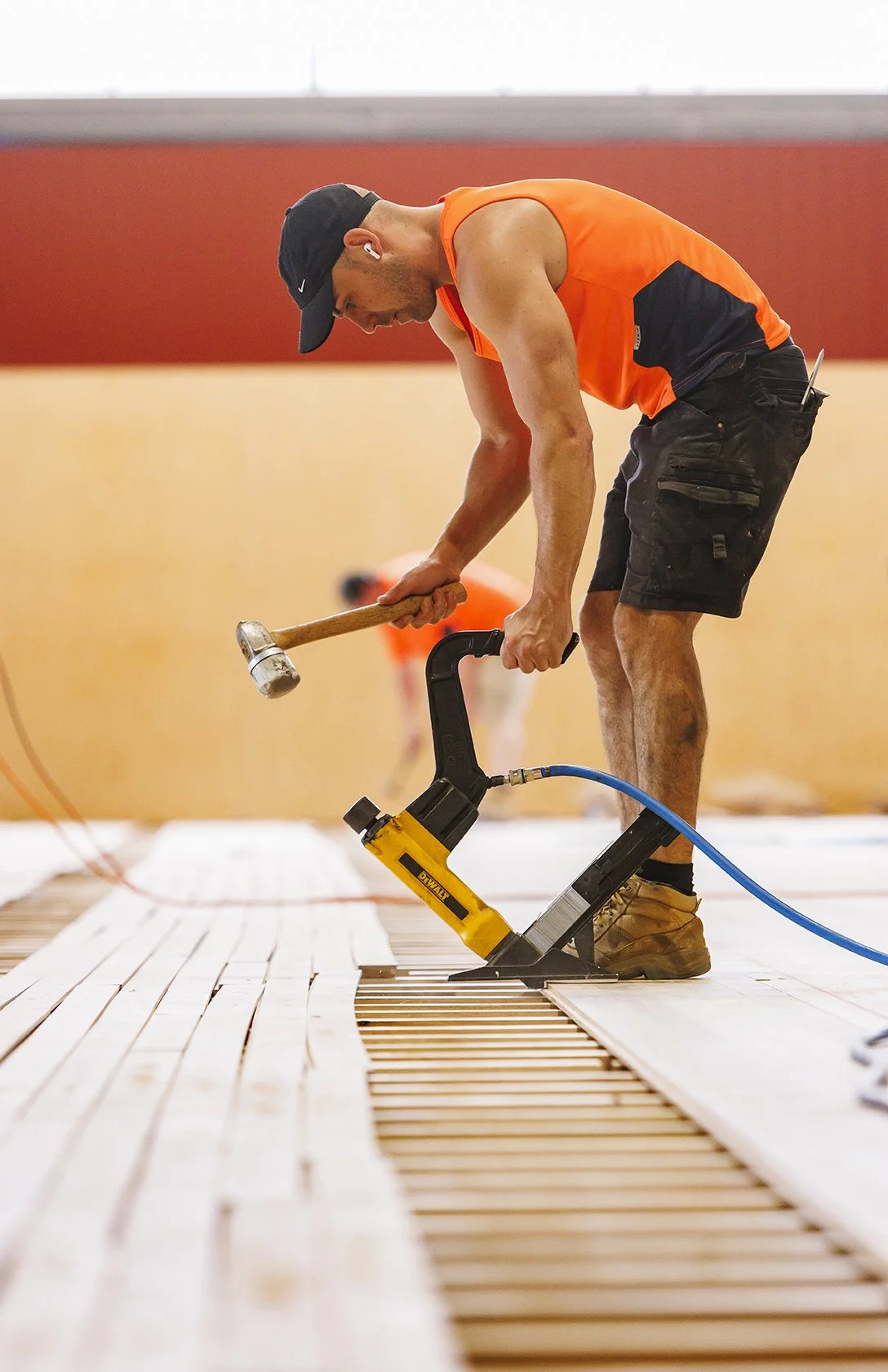 A worker in an orange tank top and black shorts using a pneumatic nail gun on wood flooring, with a hammer in hand.