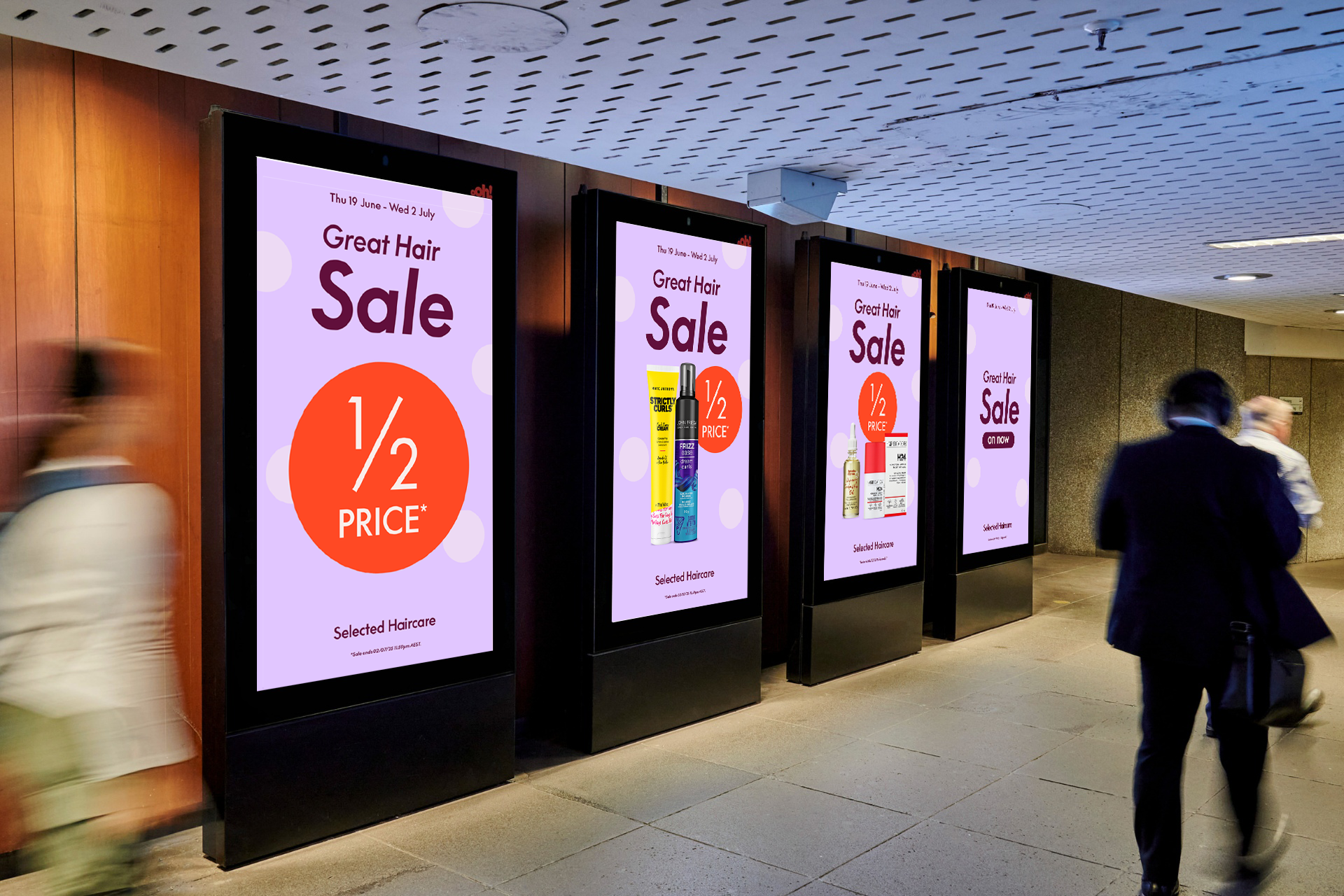 Digital advertising screens in an indoor public space promoting a haircare sale with half-price discounts, featuring different haircare products on each screen, with walking people in the foreground.
