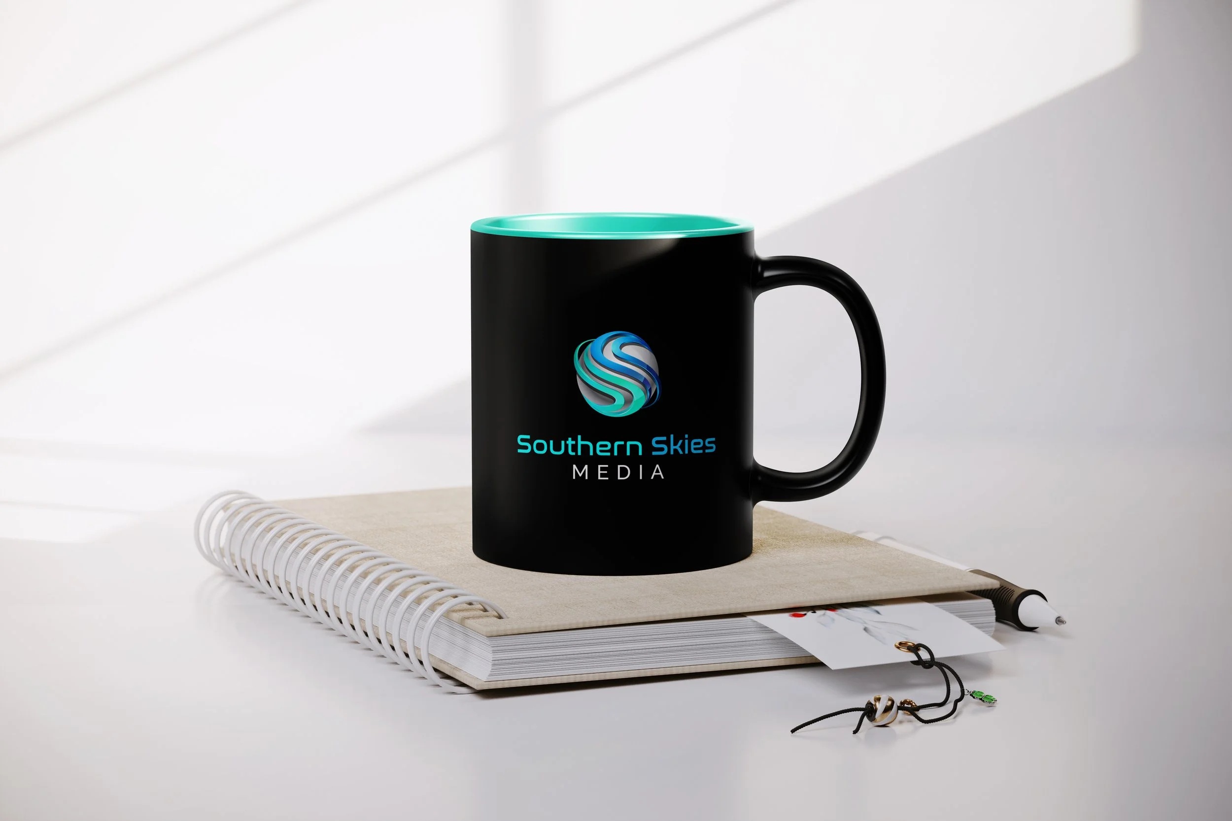 Black coffee mug with the Southern Skies Media logo and text on a desk, alongside a spiral notebook, a white pen, and a black cord with a small green keychain.