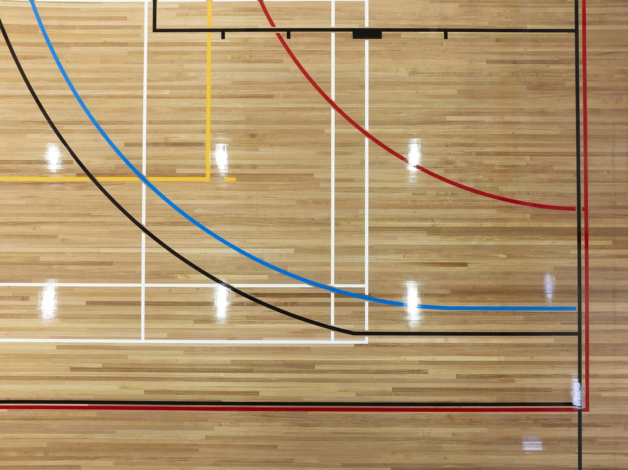 View of a basketball court from above showing boundary lines and three-point arc painted in multiple colors, including black, blue, yellow, white, and red, on a hardwood floor.