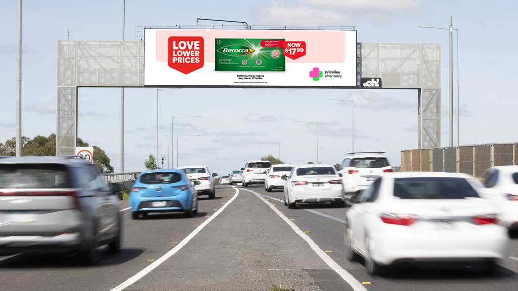 Multiple cars traveling on a highway beneath an overhead billboard displaying advertisements for a pharmacy and a low prices message, with a partly cloudy sky in the background.
