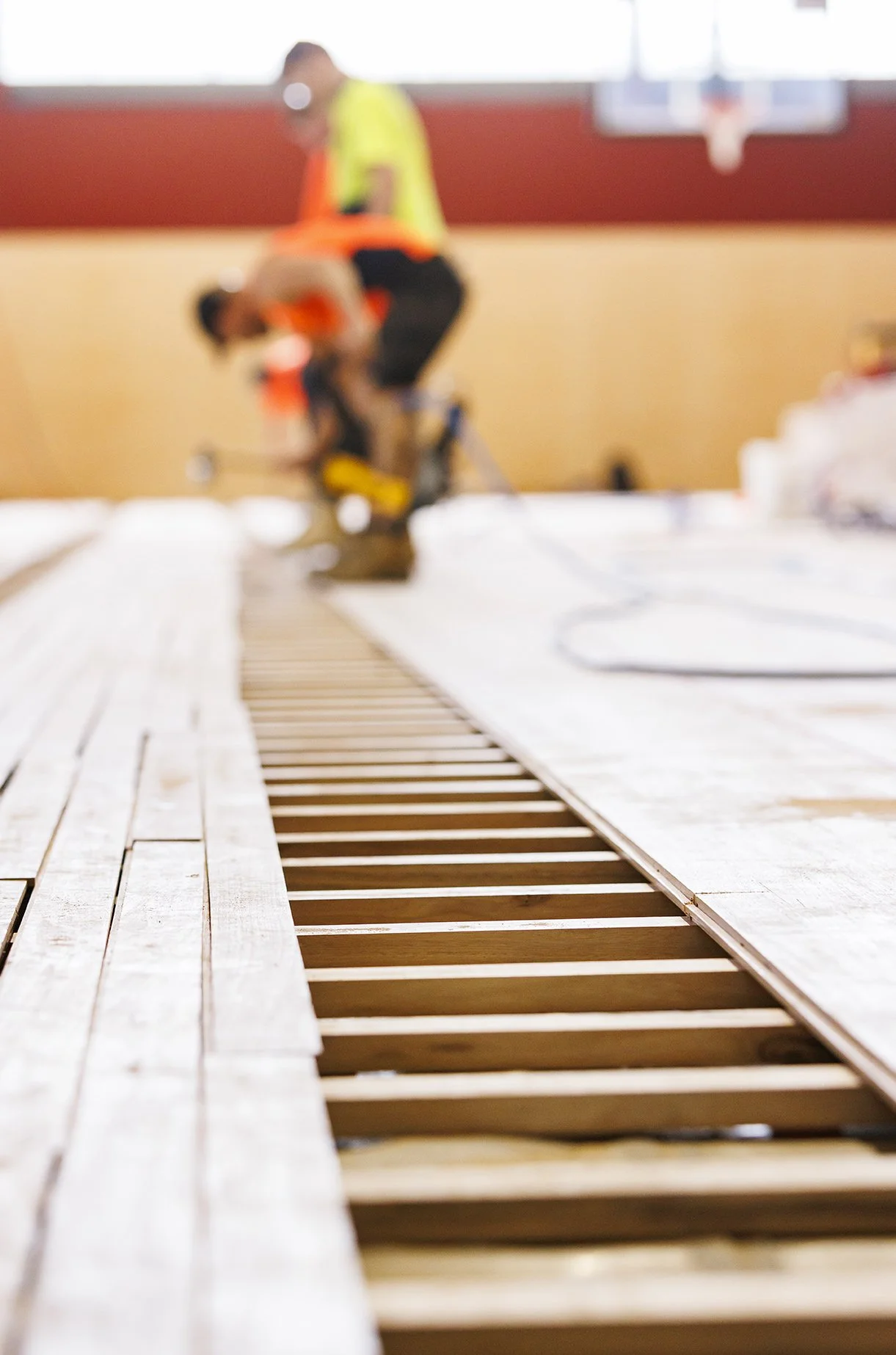Construction workers building a basketball court inside a gymnasium with a visible basketball hoop in the background.